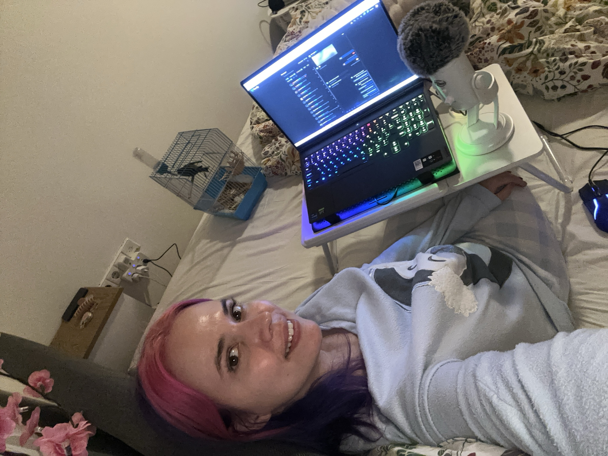 A woman with pink and purple hair is lying in bed, smiling at the camera. She is wearing a light blue sweatshirt with a dark gray logo. A laptop is on a white foldable table in front of her, with a microphone attached. The laptop screen displays a window with lines of code or a software interface. To the left of the laptop is a small birdcage with a bird inside. To the left of the woman's head is a bedside table with a power strip and a few decorative items. Pink flowers are in the foreground. The bed has a floral patterned duvet.
The location is in a residential setting, presumed to be in the City of Zagreb, Croatia.