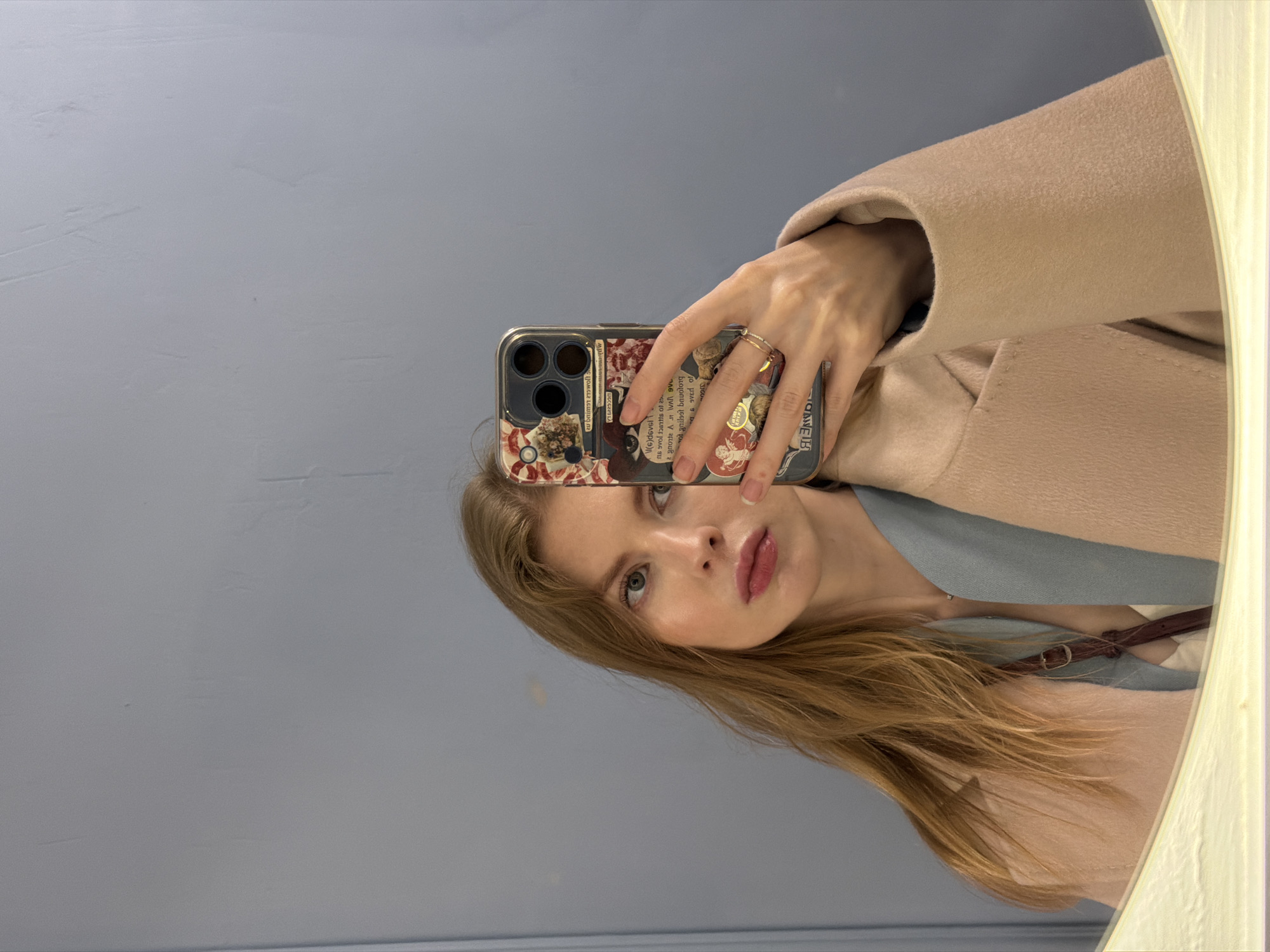 Here is a description of the image:
A woman is taking a selfie in a mirror. She is holding her phone in her right hand, the phone is patterned with a floral design. She is wearing a beige coat and a light blue top. She has long blonde hair and is looking at the camera. The background appears to be a grey wall. The location is not discernible from the provided image.