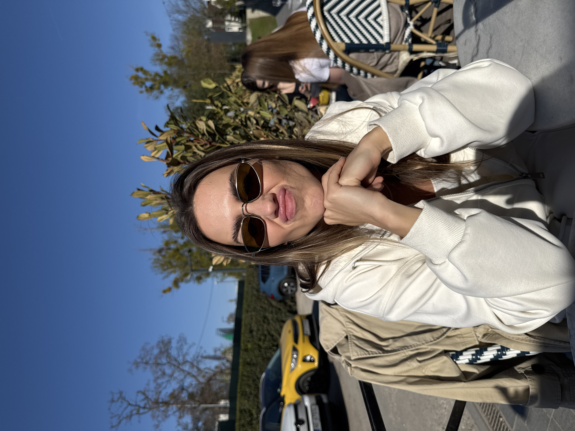 An outdoor daytime photograph taken in Warsaw, Poland, features a young woman posing playfully at what appears to be an outdoor cafe or restaurant. The main subject, with long brown hair, wears dark-lensed sunglasses and a white hooded sweatshirt. She makes a puckered lip expression while resting her chin on her clasped hands. Her right arm and shoulder are visible, with sunlight casting shadows on her.
She is seated at a table, with a beige jacket draped next to her. To her right, a black and white patterned chair, possibly woven, is partially visible. In the mid-ground, behind lush green foliage and trees, another person with long brown hair is partially seen, also seated at an outdoor table. Beyond the greenery, a street or parking area is visible with several cars, including a prominent bright yellow car (possibly a sports car) and a blue car.
The weather is sunny with a clear blue sky, casting bright light and distinct shadows, suggesting it is mid-morning or afternoon. The scene conveys a relaxed and casual atmosphere. No discernible text is visible.