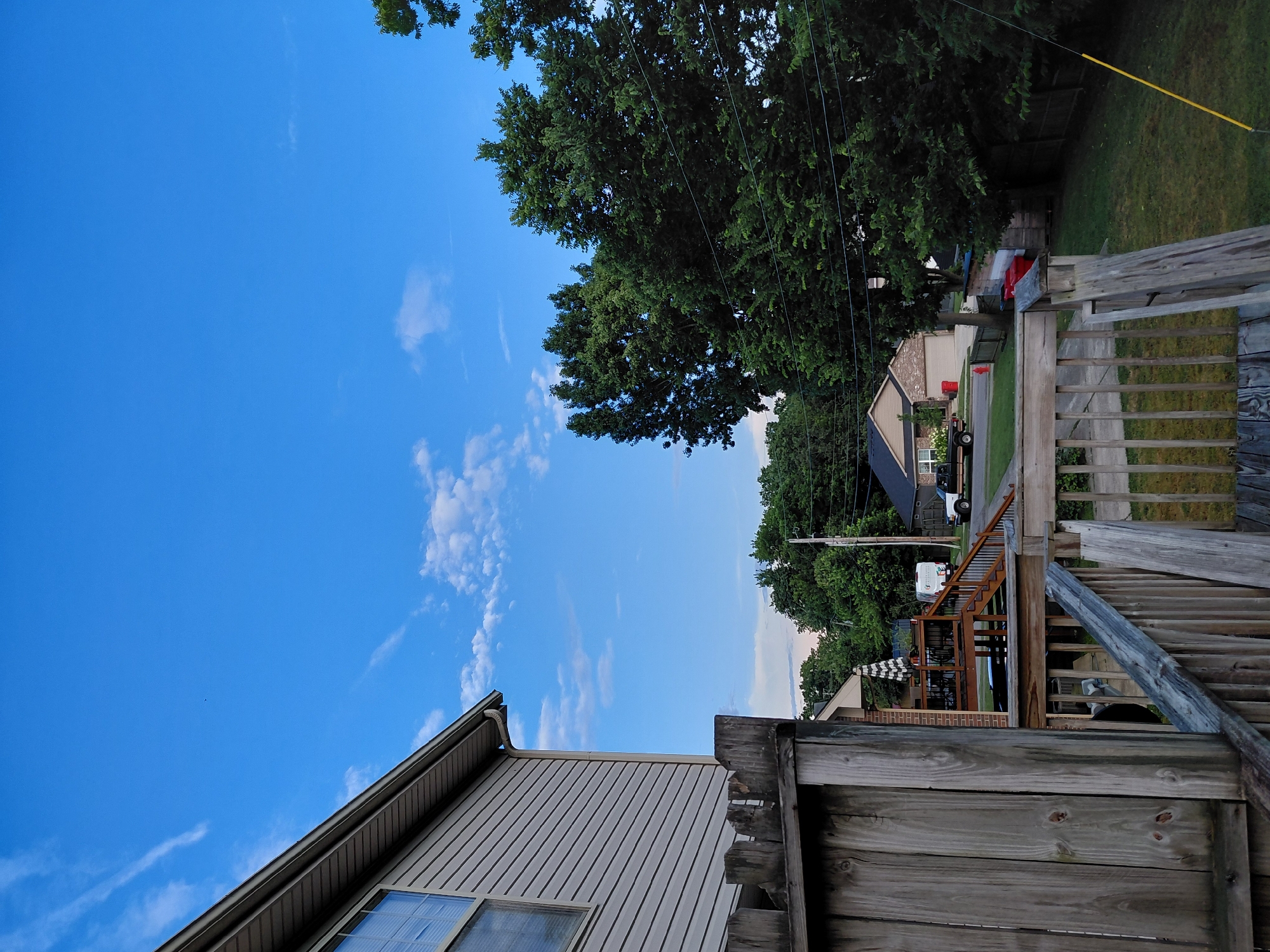 This is a medium-angle, slightly low shot taken outdoors in what appears to be a suburban residential area, likely Nicholasville, United States of America. The foreground is dominated by a weathered wooden deck and fence, with vertical wooden planks and railings. To the left, the siding of a house is visible, with a window and a portion of the roof. The middle ground features a two-story house with a deck, an umbrella, a vehicle, and a white van. Across the street, there is another house with a garage, a pickup truck, and red trash cans. Dense green trees form a backdrop on the right side of the image, with power lines strung between them. The sky is a clear blue with scattered wispy white clouds. The overall impression is one of a typical residential neighborhood during the daytime.