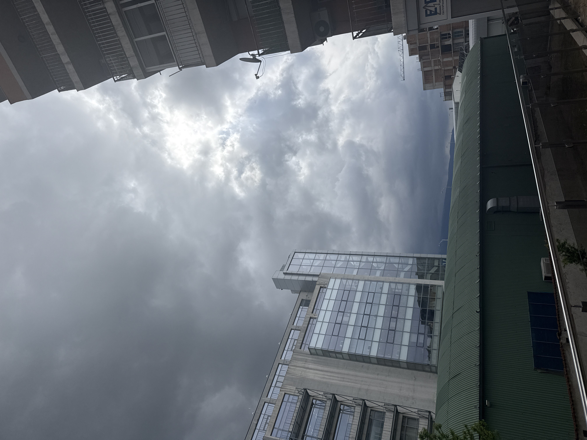 The image shows a low-angle view of a city street in Sofia, Bulgaria, under a stormy, overcast sky. The composition is framed by buildings on either side, creating a narrow perspective upward. On the left is a residential building with balconies. On the right, there's a green, corrugated building with a vertical white stripe, and a modern glass and concrete high-rise building. In the background, construction is visible with a building under construction and a crane. The sky is filled with dark, ominous clouds.