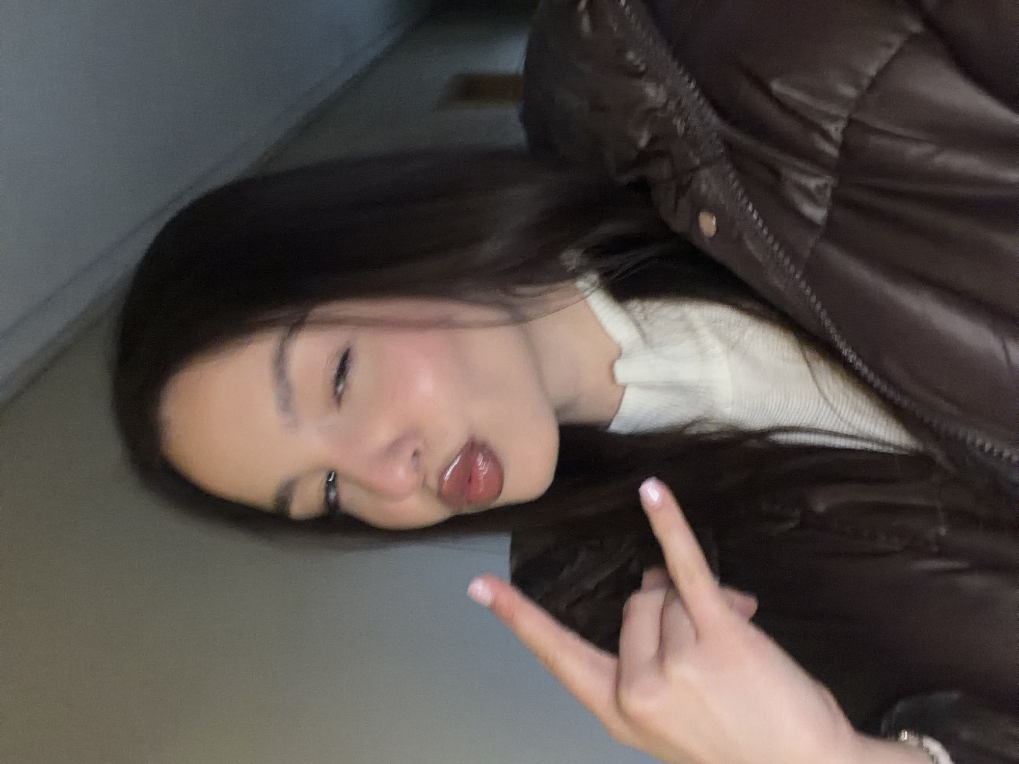 A medium shot shows a fair-skinned person with long, dark hair, wearing a white top and a dark, puffy jacket. The person's face is angled with a pouting expression, and they are extending their middle finger in front of them. The background appears to be an interior space with neutral-colored walls. The photograph is slightly out of focus. The location is not discernible from the image.