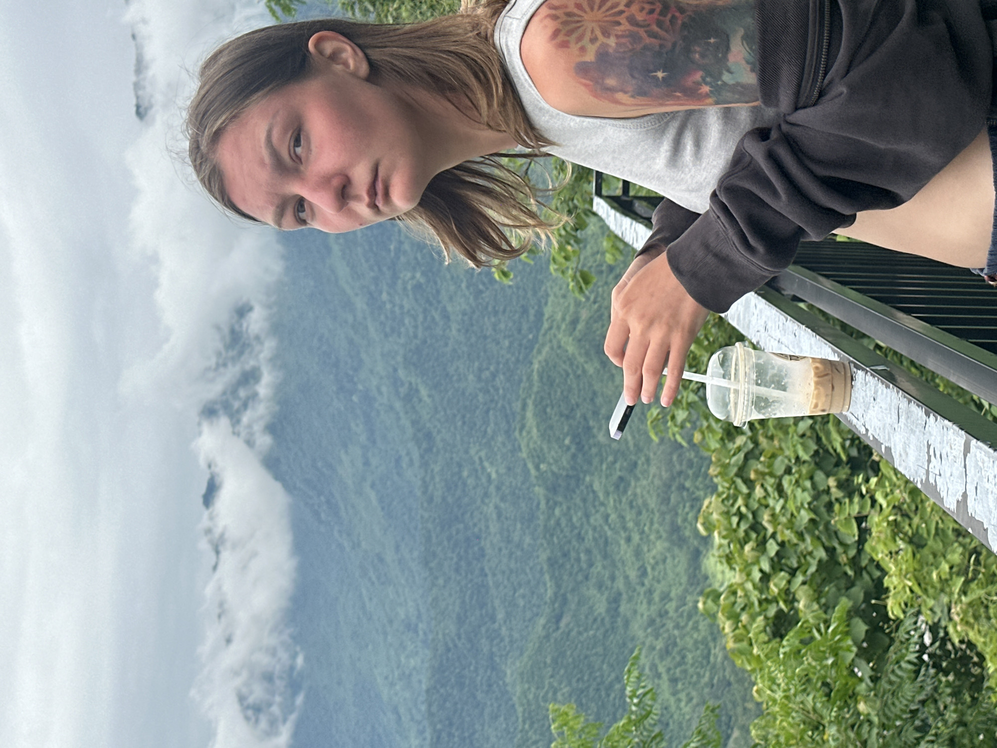 A young woman with light brown hair stands on a balcony overlooking a lush green mountain landscape under a cloudy sky. She is wearing a grey tank top and a dark grey zip-up hoodie. Her left arm, which is visible, features a colorful tattoo of a celestial scene with stars and a sunburst-like design. She is holding a smartphone in her right hand and a clear plastic cup with a straw, containing a light brown beverage, is placed on the balcony railing in front of her. The railing is made of metal and appears weathered. The background is dominated by densely forested mountains, partially obscured by mist and clouds. This scene is characteristic of viewpoints found in Mai Chau, Vietnam, known for its mountainous terrain and misty conditions.