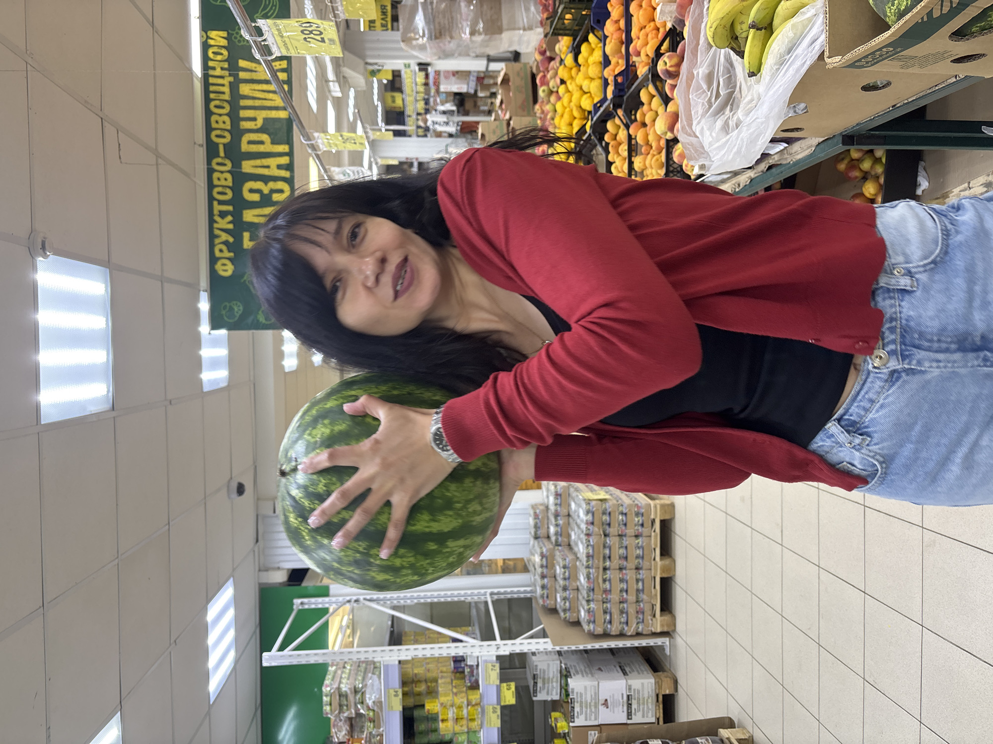 The image, rotated 90 degrees clockwise, captures a woman in a supermarket or grocery store, likely within the produce section. The woman, with dark hair, wears a red cardigan over a black top and blue jeans, with a silver watch on her left wrist. She holds a large green watermelon with both hands and smiles towards the camera.
The background shows store aisles stocked with various fruits, including bananas, peaches, and apricots in crates and boxes. Shelves with other packaged goods are also visible. The store interior features white tiled floors, bright rectangular overhead lights, and a security camera mounted on the wall.
A prominent green sign above the produce section displays Cyrillic text, reading "ФРУКТОВО-ОВОЩНОЙ БАЗАРЧИК," which translates to "FRUIT-VEGETABLE MARKET/BAZAAR." Several yellow price tags are visible, showing numbers such as "289," "74," and "99." The overall scene depicts a casual shopping experience.