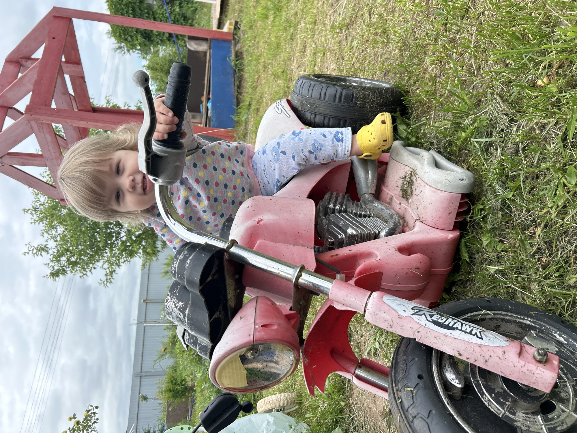 A blonde-haired toddler sits on a pink toy motorcycle in a grassy outdoor area. The child is wearing a grey polka-dot shirt and light blue pants with a rabbit pattern, and yellow Crocs. The motorcycle has a prominent front headlight, a black seat, and a silver handlebar. In the background, a partially constructed red wooden structure is visible, along with a gray metal fence and greenery. The scene appears to be in a residential backyard or a play area in the village of Olkhovka, Russia.