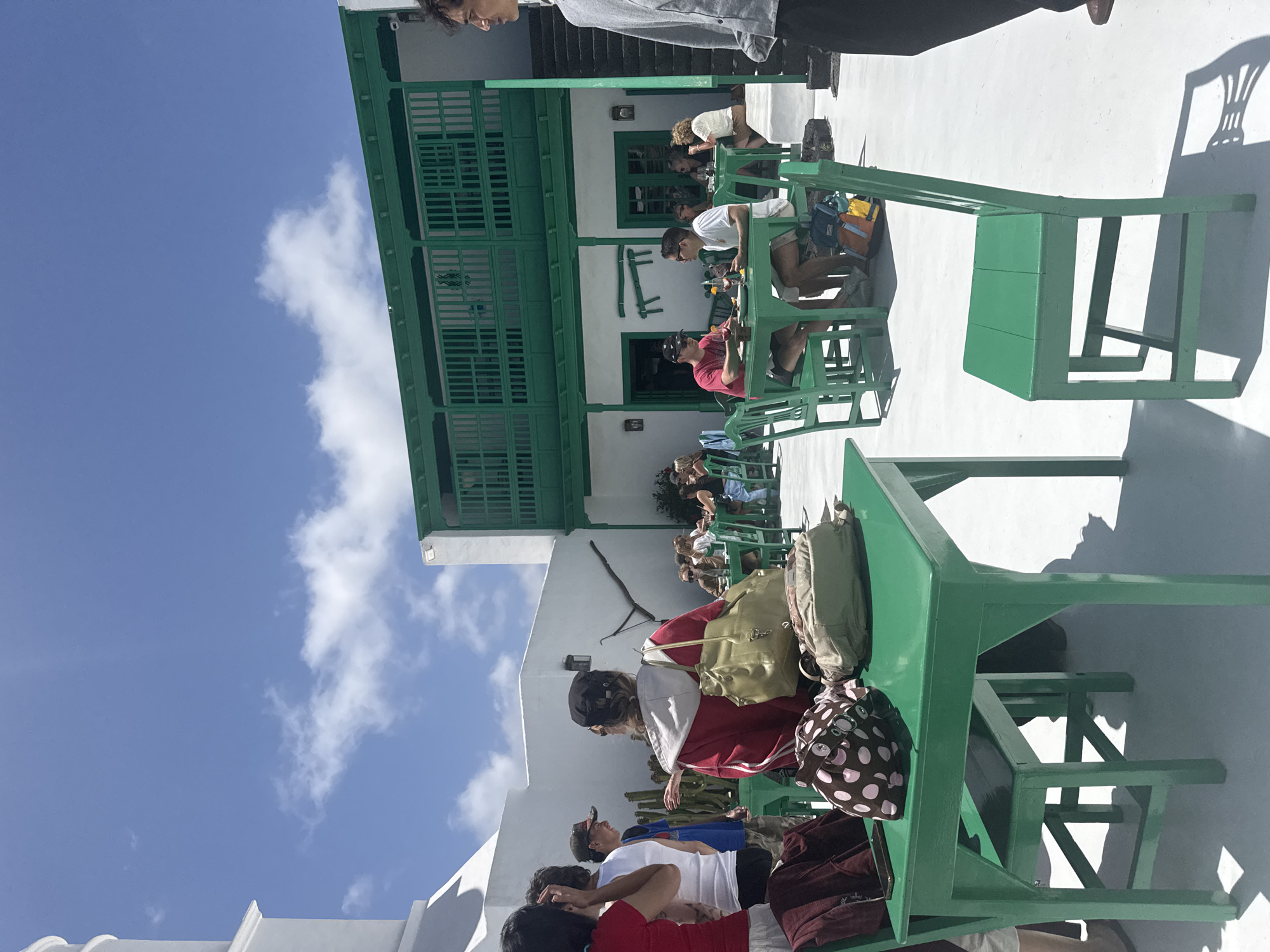 The image shows a bright, sunny day at Casa Museo del Campesino, a museum located in San Bartolomé, Spain. Several people are seated at bright green tables and chairs, presumably tourists or visitors. Some appear to be resting or eating, while others are looking around. The building behind them is white with green trim around the windows and a green balcony. A few white clouds dot the blue sky. Backpacks and other personal belongings are placed on the tables.