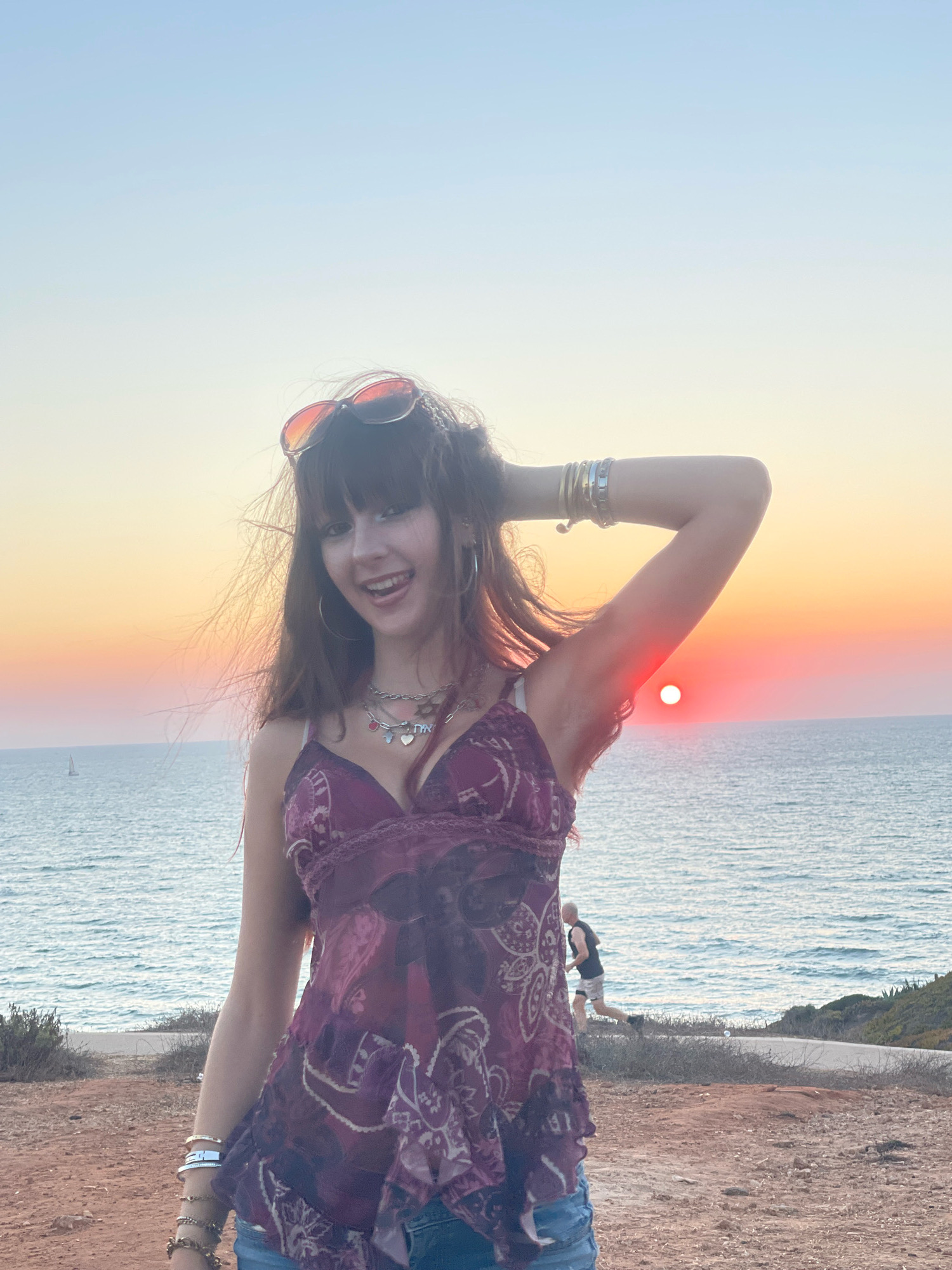 A young woman with long brown hair and bangs, wearing a purple floral print top, denim shorts, sunglasses, and several bracelets, stands on a reddish-brown, dirt path overlooking the Mediterranean Sea at sunset. She is smiling and has her right arm raised behind her head. A bright orange sun is visible on the horizon, casting a warm glow across the sky and water. In the background, a man is jogging along a path near the coastline, and a sailboat is visible in the distance. The scene appears to be from a coastal area in Herzliya, Israel.