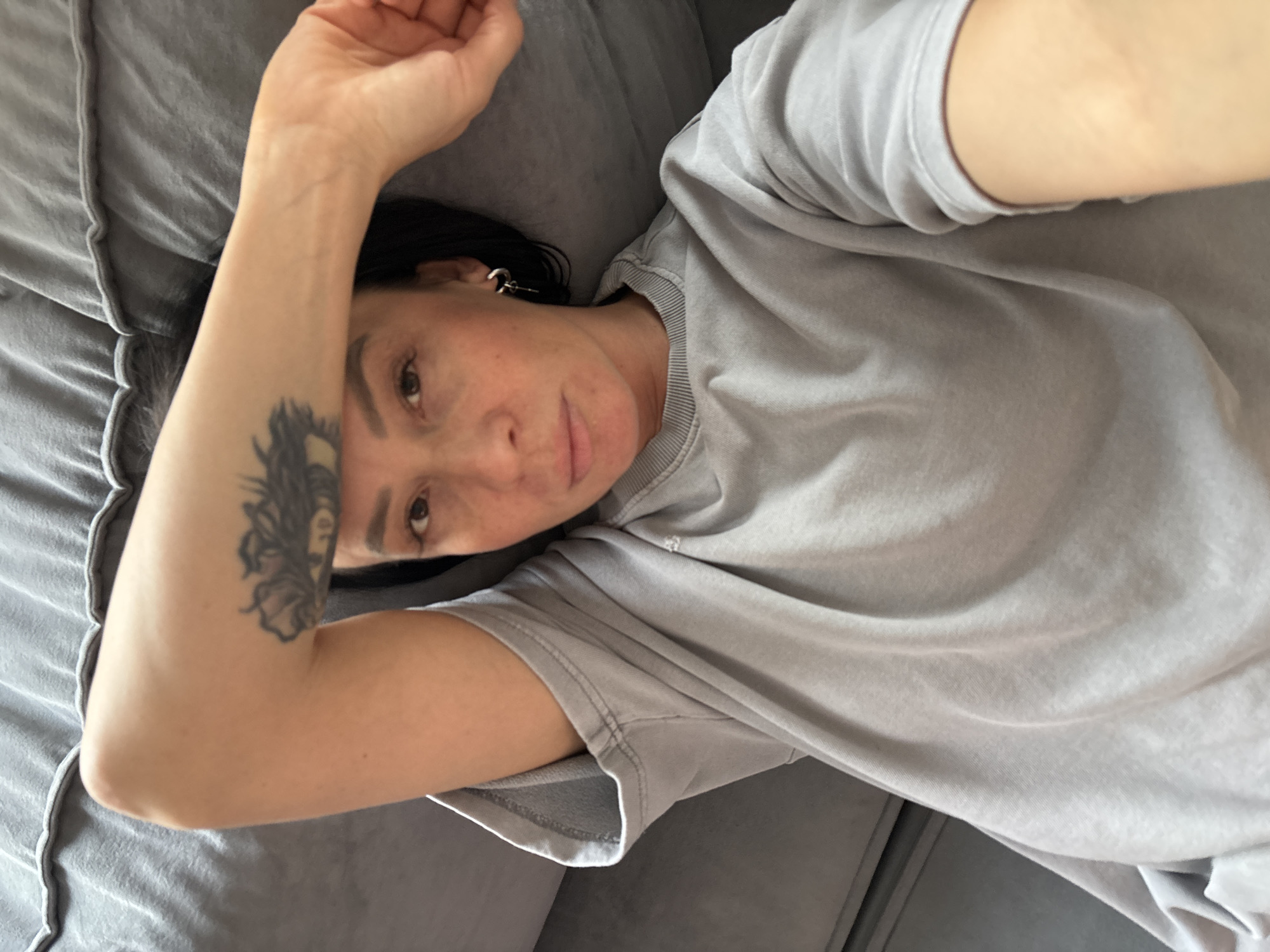 An overhead photograph depicts a female individual lying supine on a textured grey upholstered surface, likely a sofa or bed. She wears a grey, short-sleeved or rolled-sleeve crew-neck garment, possibly a sweatshirt or t-shirt, with visible stitching details. Her right arm is bent, with the forearm positioned horizontally across her upper face, partially obscuring her forehead. A dark-inked tattoo, resembling a stylized eye or a feathered pattern, is visible on the exterior of her right forearm. Her dark hair is visible on the left side of her head. A silver-colored hoop earring with a small, dark dangling element is present in her left ear. Her eyes are open and gaze directly towards the camera. The skin on her face, particularly her cheeks and chin, exhibits minor redness. The specific location within the city of Chaikovsky, Russia, cannot be determined from the visual information provided in the image.