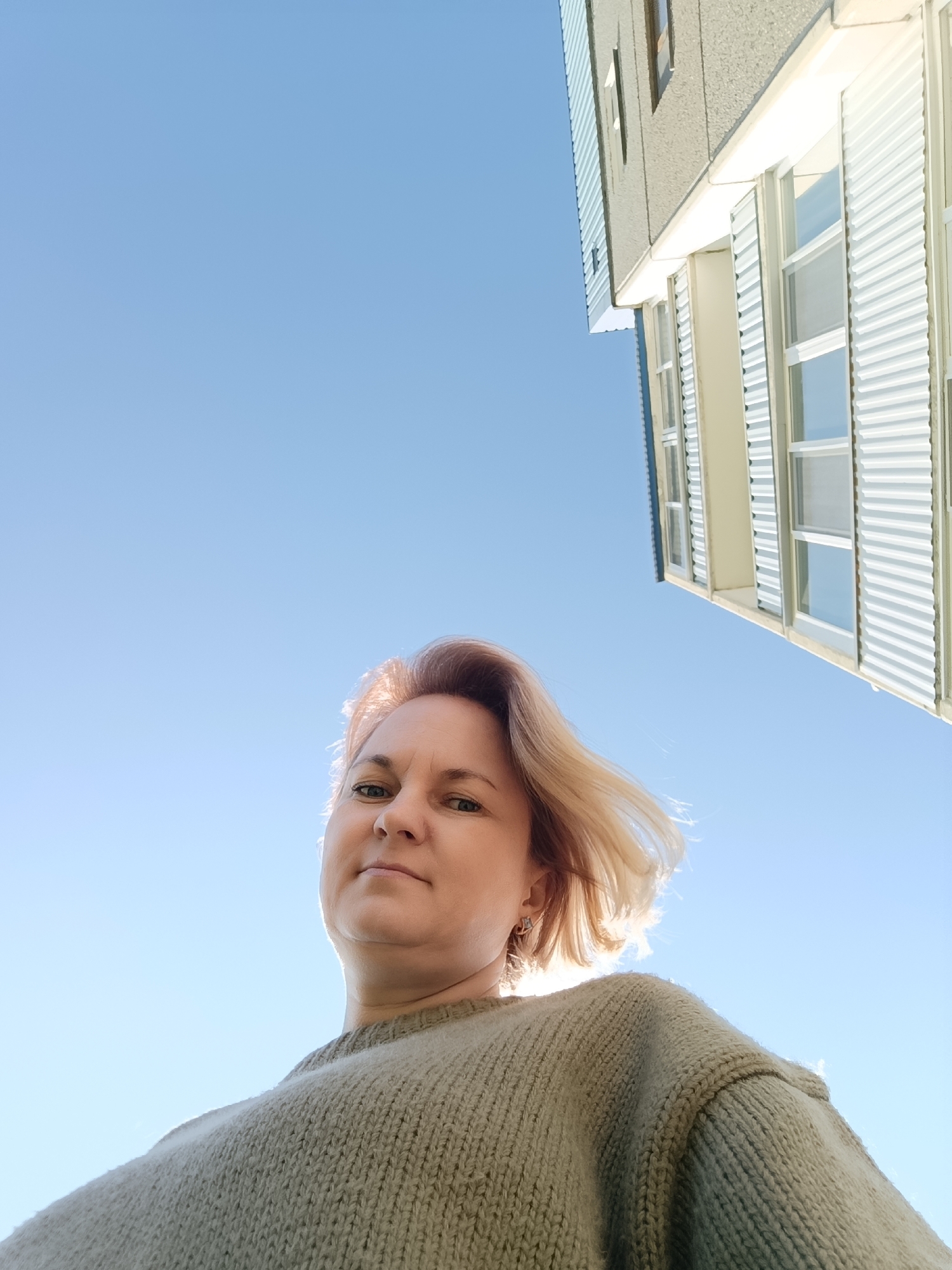 A low-angle outdoor shot features a woman looking directly up at the camera, set against a bright blue sky and a portion of a building.
The woman occupies the lower center of the frame, gazing forward with a neutral to slightly serene expression. She has fair skin and light-colored hair, possibly blonde or light brown, which is backlit by strong sunlight, creating a luminous glow around her head and appearing gently windswept. She is wearing a thick, chunky knit sweater in a muted olive green or light green color, which takes up a significant part of the lower frame.
The upper two-thirds of the image showcase a vast, cloudless, and vibrant blue sky, indicating clear and sunny weather. In the upper right corner, a section of a multi-story building is visible, angled diagonally. The building features a textured, possibly concrete or stucco facade, and prominent windows with white horizontal slats or louvers, suggesting a contemporary architectural style. The sunlight strongly illuminates the building's exterior.
The scene conveys a bright, sunny daytime setting, likely morning or afternoon, given the intense backlighting on the woman and the general illumination. No text is visible in the image. The environment is an outdoor urban or suburban landscape, situated in the Марковский, Russia context.