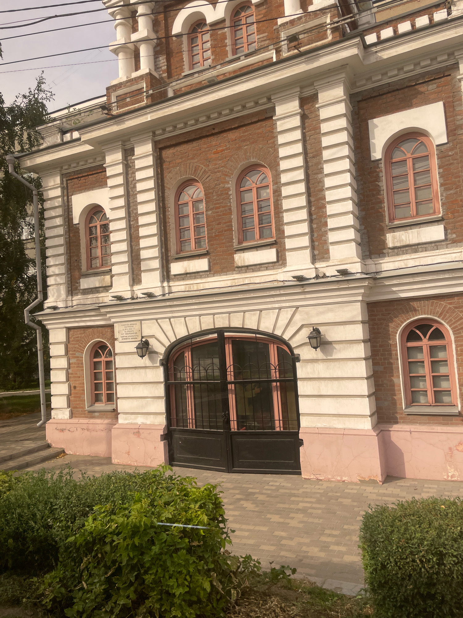 The image shows the facade of a brick building in Omsk, Russia. The building features a classical architectural style with arched windows, pilasters, and decorative cornices. The windows have light pink frames and mullions. A large black metal gate with pink accents is centrally located on the ground floor, beneath a prominent arched entryway. A small sign with text in Cyrillic is mounted on the wall above a black lamp fixture. In the foreground, there is a well-maintained green bush. The ground is paved with interlocking rectangular stones. The sky is overcast.