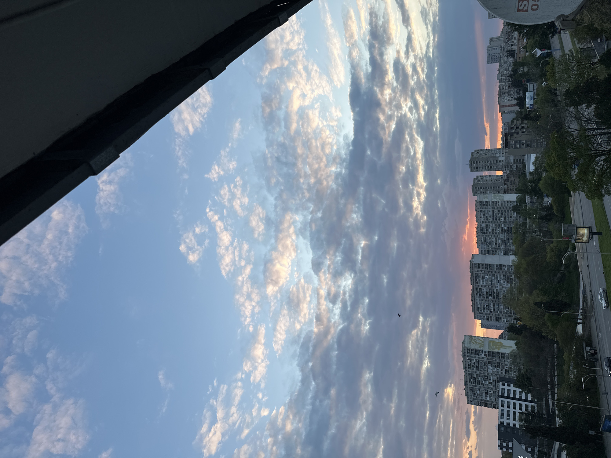 The image displays an urban landscape at either sunrise or sunset, viewed from an elevated perspective, likely a balcony or building. The sky dominates the upper portion, filled with layered altocumulus or stratocumulus clouds. The clouds near the horizon glow with warm hues of orange, pink, and yellow, blending into cooler blues and greys higher up, indicating the soft light of dusk or dawn. Patches of clear blue sky are visible between the clouds.
In the lower half of the image, a cityscape unfolds. Numerous multi-story apartment buildings, appearing to be concrete construction, are densely packed. One prominent building features a large, light-colored mural or graffiti on its facade. A road runs through the scene, with vehicles visible, suggesting ongoing activity. Trees and greenery are scattered between the buildings and alongside the road, providing some natural elements within the urban environment.
Several small, dark bird silhouettes are visible against the illuminated sky, adding life to the serene atmosphere. A dark, architectural overhang from the vantage point partially frames the top-left corner of the image. On the far right, a white satellite dish is mounted, with the letters "S" and numbers "05" visible on its surface. The overall mood is peaceful and contemplative, capturing the quiet beauty of a transitional time of day in a bustling city.