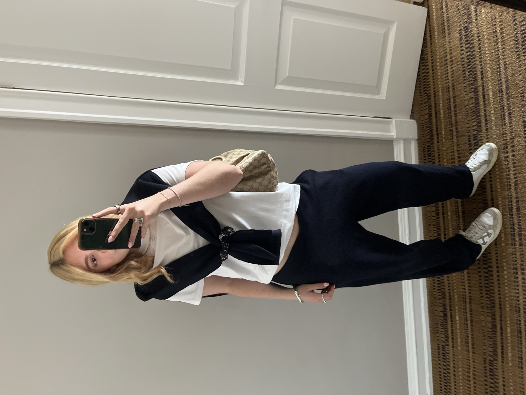 A person is taking a mirror selfie in what appears to be a room with a door and a carpeted floor. The person has blond hair and is wearing a white T-shirt with a dark blue sweater tied around the waist, along with dark blue pants and white sneakers. They are holding a phone with a dark green case. A small, patterned handbag is slung over their shoulder. The wall is a light gray color, and the door is white. The carpet has a brown and gold patterned design. The setting does not definitively indicate Johannesburg, South Africa.