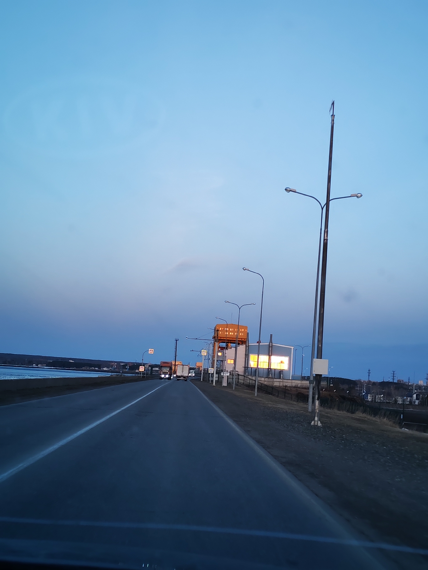 The image depicts a road in Chaikovsky, Russia at dusk. The road stretches into the distance with a white dividing line, and several trucks are visible further down the road. On the right side of the road, there are a series of streetlights and buildings. Behind the buildings, the city can be seen in the distance. On the left side of the road, there is a body of water, likely a river or lake. The sky is a gradient of blue and purple, indicating the time of day. Road signs are present on the side of the road.