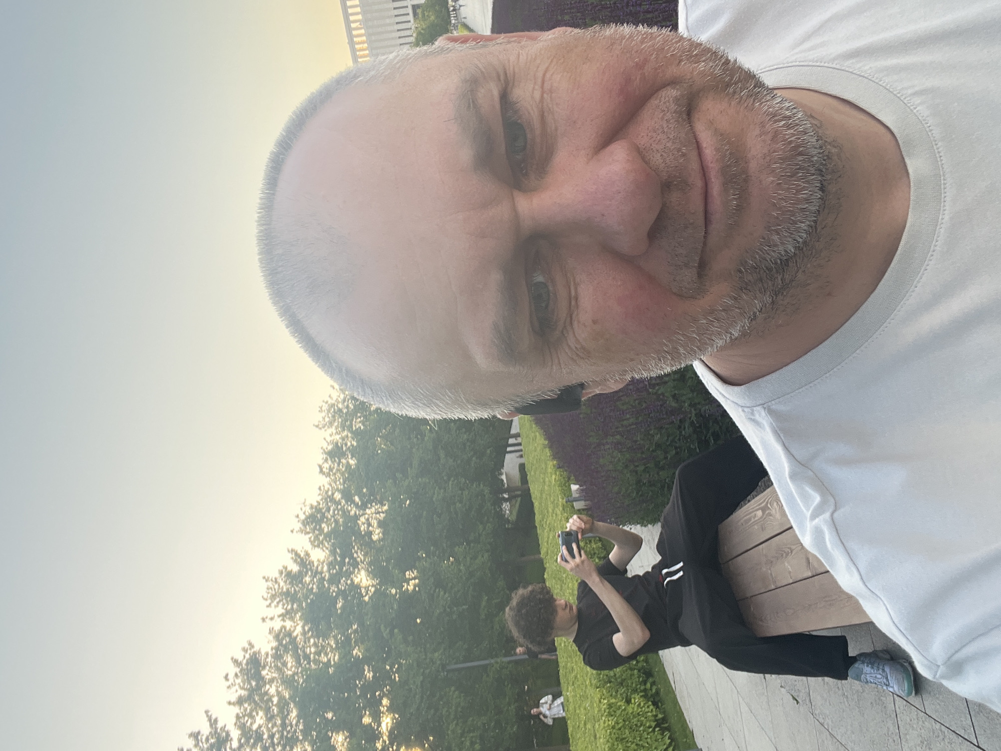 A selfie depicts a man with grey hair and beard wearing a white t-shirt. In the background, a younger person sits on a bench and takes a photograph with a mobile phone. The scene includes greenery and a building facade partially visible at the top right. The location is suggested to be in Krasnodar, Russia.