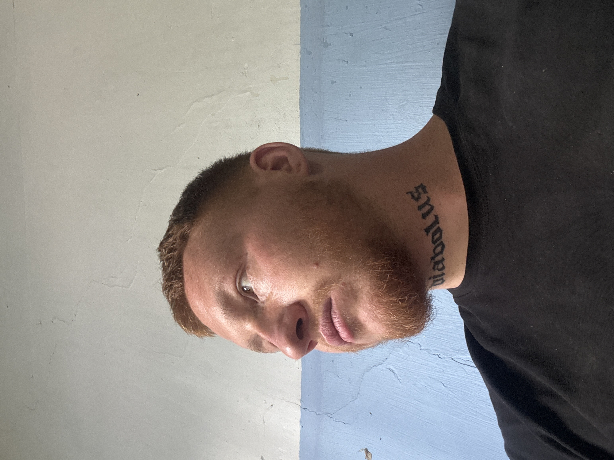 A close-up, upward-facing shot shows a fair-skinned, reddish-haired man with a reddish beard, wearing a black shirt. He has a tattoo on the right side of his neck written in a gothic font. The background consists of a wall painted white on one side and blue on the other. There is no information to suggest the photo was taken in the city of Makiivka, Ukraine.