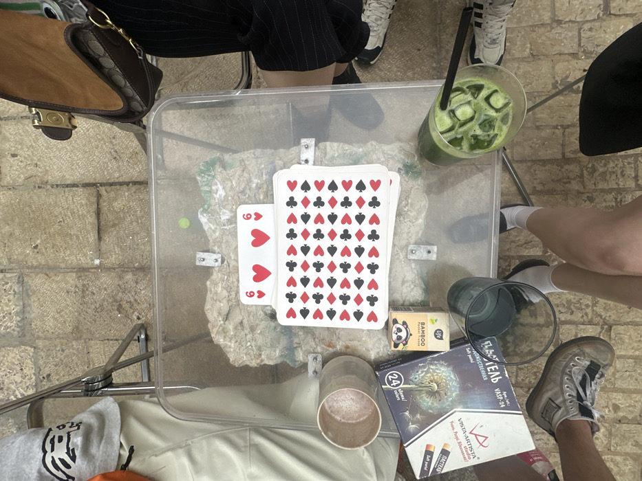 An overhead view captures an outdoor scene in Tel Aviv-Yafo, Israel, featuring a clear, rectangular table situated on a light-colored stone-tiled surface. The table's base consists of exposed rock or concrete, secured by metal brackets visible through the transparent top. Central on the table is a large stack of playing cards; the top card displays the 6 of Hearts, while the visible backs of the other cards feature a repeating pattern of red hearts, red diamonds, black clubs, and black spades.
Adjacent to the cards are a light brown ceramic mug, a rectangular box of "VISTA-ARTISTA VASP-24 soft pastel" depicting a dandelion, and a small "BAMBOO Pocket Tissue LIFE" box with a panda logo. A tall glass containing a green iced beverage with a black straw is present, alongside another empty glass.
Around the table, parts of at least three individuals are visible:
To the left, a person wears beige pants and grey sneakers, with a grey sweatshirt displaying a black graphic.
To the right, a person wears dark pinstriped pants and white Adidas sneakers, with a brown suede shoulder bag featuring a patterned strap and a gold buckle.
Towards the bottom-center, a person's bare legs, white socks, and white sneakers are visible, with dark clothing (possibly shorts or a skirt) below the knee. The scene suggests a casual gathering.