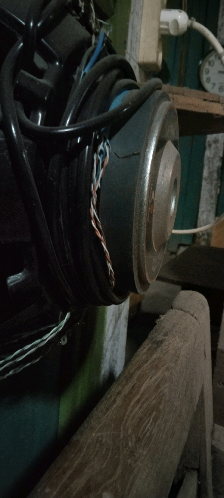 The image depicts a close-up, interior shot of what appears to be a piece of electrical equipment, possibly a speaker, with various wires and cables connected to it. The main subject is the metallic rim of the speaker cone, which has a ribbed texture. A cluster of thin, multi-colored wires (white, orange, blue) are twisted together and emerge from the black casing of the speaker. Black, thick cables are also visible, coiled around the speaker.
In the background, a white electrical outlet is plugged into a light-colored wooden surface. To the right of the outlet, a circular clock with visible numerals (8, 9, 10, 11, 12, 1, 2, 3, 4, 5, 6, 7) is partially visible. A rustic wooden beam with a rough texture runs diagonally across the foreground, obscuring a portion of the speaker and the background. The environment suggests an indoor, possibly storage or workshop setting, with a rustic aesthetic. There are no people or clear indications of specific actions or interactions. The lighting is dim, contributing to a shadowed and somewhat cluttered appearance. No specific location in Wadang, Indonesia can be determined from the visual information provided in the image.