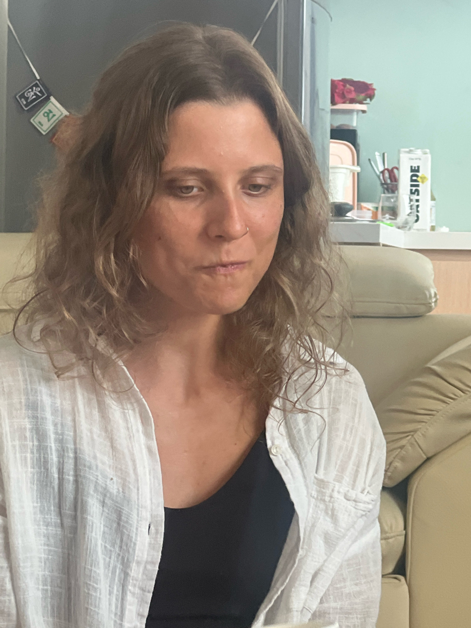 A medium shot captures a woman with shoulder-length wavy brown hair, looking down and slightly to her left. She is wearing a white button-up shirt with a black tank top underneath and a nose ring. Her expression is neutral. The background includes a beige sofa, a kitchen counter with various items, and a light blue wall. In the background, on the left, there is a refrigerator door with two small square stickers. On the right, there is a carton with the text "OUTSIDE" and some utensils in a holder. The overall scene suggests an indoor domestic setting, possibly an apartment, in Đà Nẵng, Vietnam.