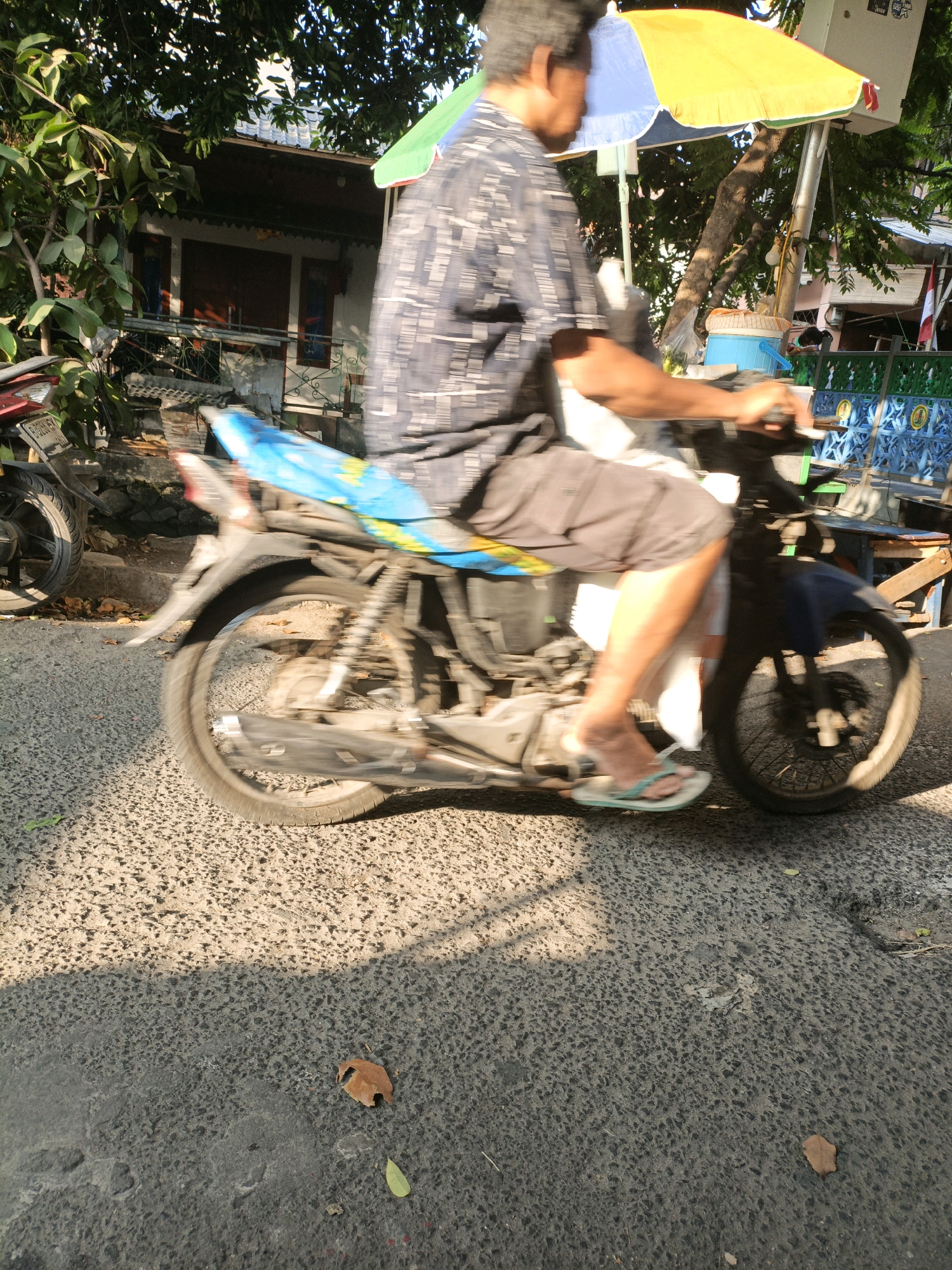 In West Jakarta, Indonesia, a man is riding a motorcycle on a paved road. He is wearing a patterned short-sleeved shirt and dark shorts, with light-colored flip-flops on his feet. The motorcycle has a blue seat cover and a dark-colored front fairing. Sunlight casts shadows on the road. In the background, there is a building with a white facade, a dark roof, and open doorways, partially obscured by trees and foliage. A yellow and blue umbrella is visible above and behind the motorcycle, and to the right, there appears to be a market stall with a blue and green fence. Another motorcycle is parked to the left of the frame.