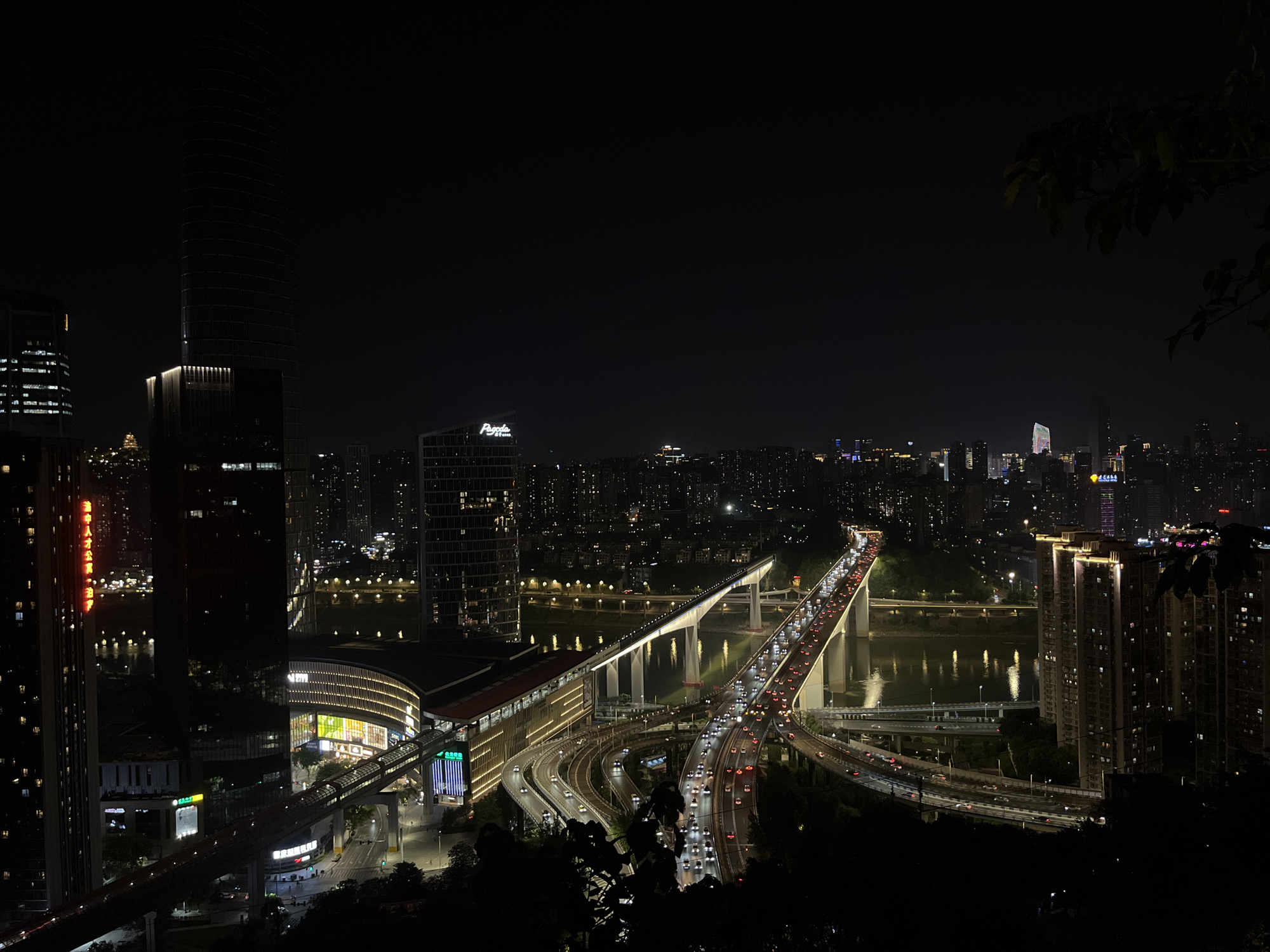 The image shows an illuminated cityscape at night, likely Yuzhong District, China. A river divides the scene. A large bridge spans the river, congested with cars. Multiple highway ramps and roadways interconnect. Buildings of varying heights are dispersed throughout the urban landscape. Some structures have illuminated facades, including a skyscraper on the left, and one with a prominent "P" logo on top. The scene is viewed from a slightly elevated position, with foliage visible in the foreground on the right.