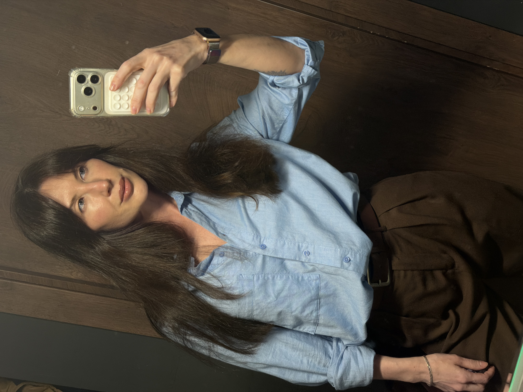 A fair-skinned woman is taking a selfie in front of a dark wood-paneled background. She is wearing a light blue collared shirt and dark brown pants. Her long, dark brown hair is partially obscuring her face. She is holding a smartphone with a protective case in her right hand, and her left hand is resting on her lap. She also wears a wristwatch on her right wrist and a bracelet on her left wrist. The location is likely indoors, possibly in an apartment or home in the city of Perm, Russia.