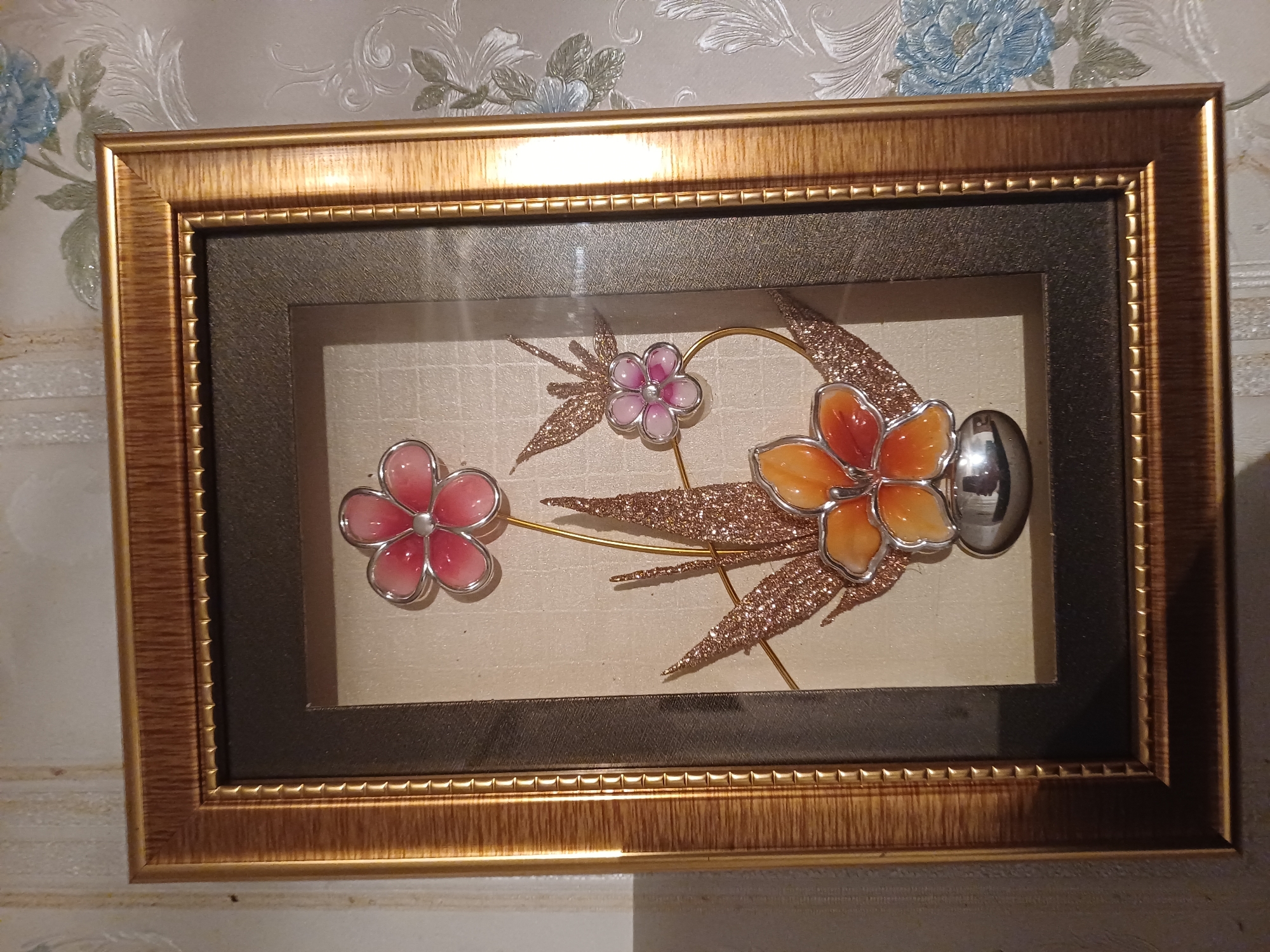 A framed artwork of artificial flowers is displayed against a textured, patterned wallpaper. The artwork features three stylized flowers with metallic silver outlines. The largest flower, at the top, has pink petals. Below it, a smaller pink flower with a darker pink center is positioned to the right. A larger orange and red flower is at the bottom center, appearing to be in a silver vase. Glittery, leaf-like elements in a rose gold color are interspersed with the flowers. The frame is golden with a ribbed texture. The wallpaper is light-colored with a repeating pattern of blue and green floral designs. The image is taken from a slightly downward angle, with the top of the frame visible.
