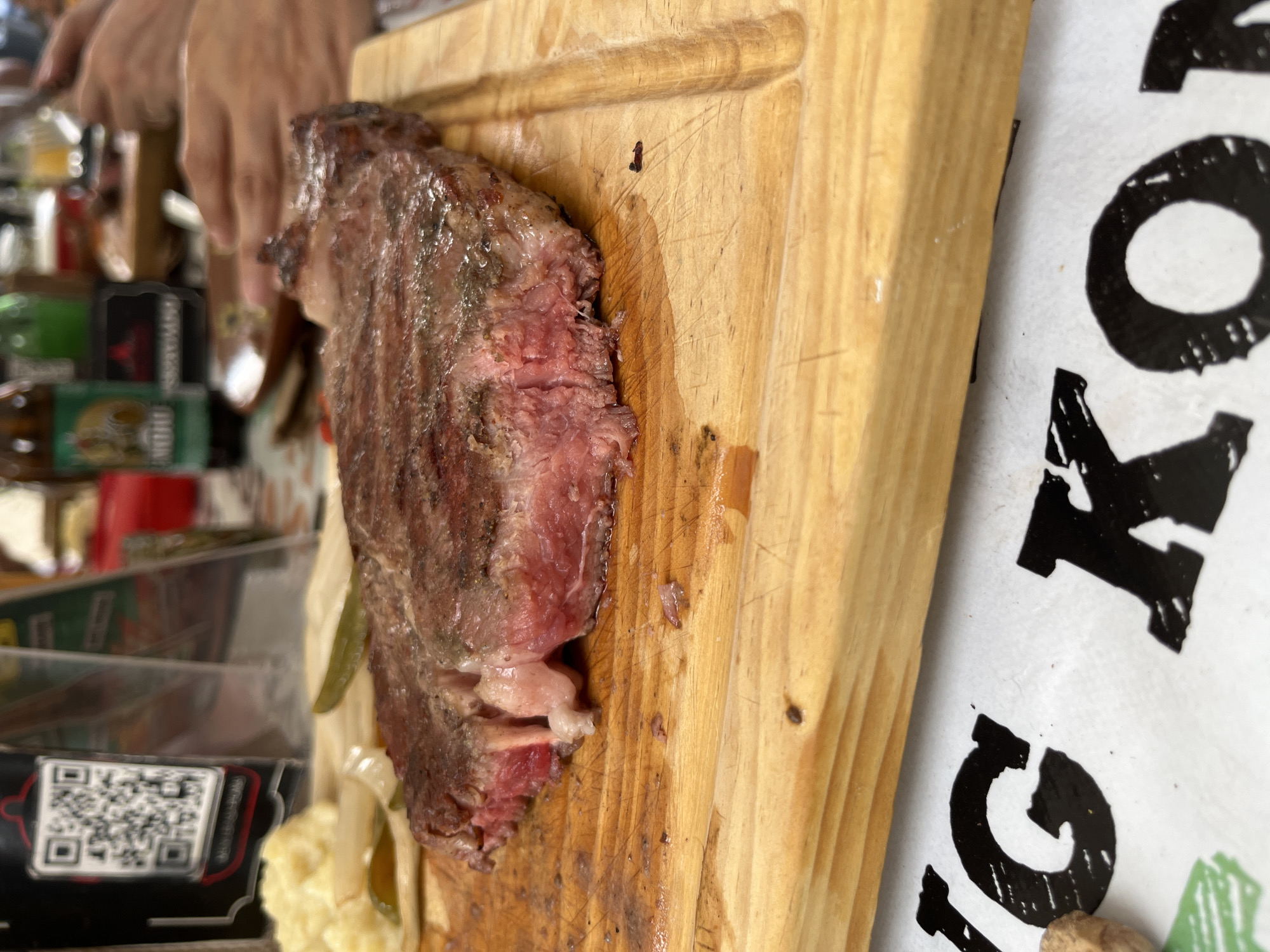 The image captures a close-up view of a piece of meat served on a wooden cutting board. The meat appears to be cooked, with a reddish-pink center and a browned exterior. It sits alongside some pickled vegetables and mashed potatoes. The wooden board shows signs of use, with knife marks and oily residue. In the background, a person's hands are visible, suggesting someone is preparing or serving the food. A QR code is also visible in the background. The environment seems to be an indoor setting, possibly a restaurant, though the specific location within San Francisco Totimehuacán, Mexico, cannot be determined from the image alone. A sign is visible with part of the word "KONG".