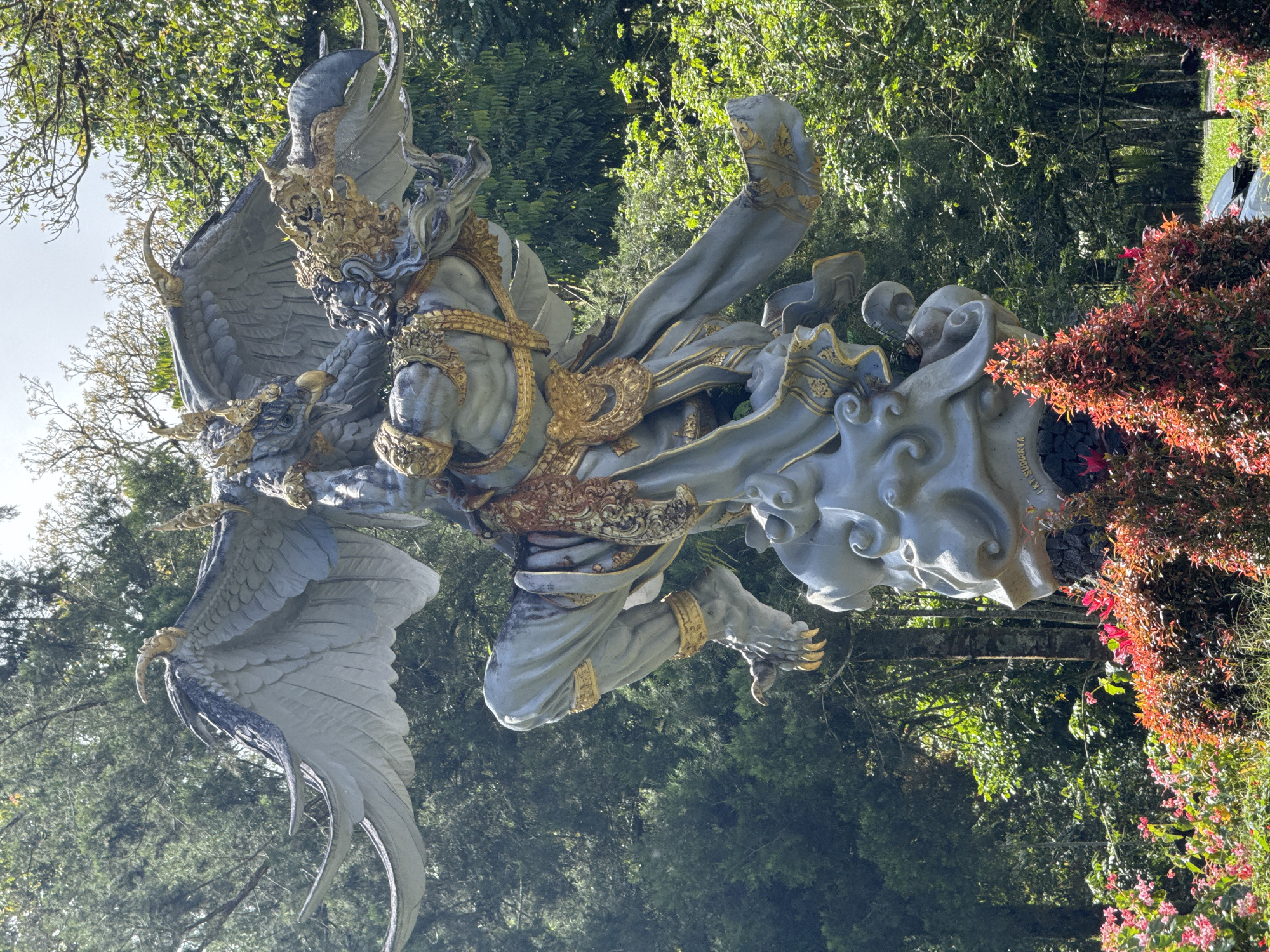 The image shows a large, intricately detailed statue of a mythical creature, possibly Garuda, in a park-like setting. The statue is predominantly light gray with gold accents on the armor and ornamentation. The creature has large wings, clawed feet, and a fierce expression. It appears to be riding or holding onto a cloud-like form. The statue is situated in a lush, green environment with trees and flowering bushes. The location is in Baturiti, Indonesia.