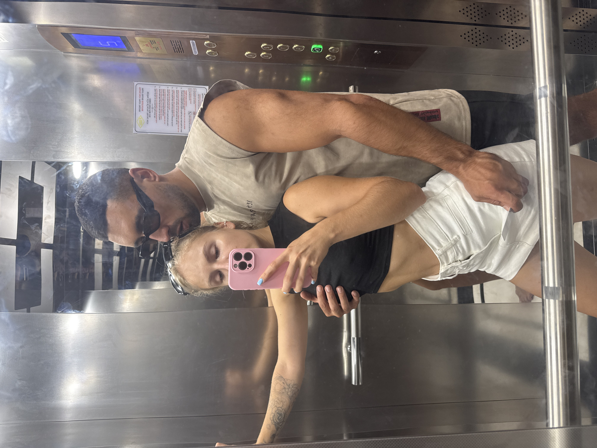 In an elevator in Nha Trang, Vietnam, a young woman with blonde hair, wearing a black crop top and white shorts, holds a pink smartphone to take a selfie. She is embraced by a man with dark hair and sunglasses, who is wearing a beige sleeveless t-shirt and black shorts. The elevator walls are made of polished metal. A digital display on the wall indicates the elevator is on the fifth floor. The scene is a candid moment captured within the confined space of the elevator.