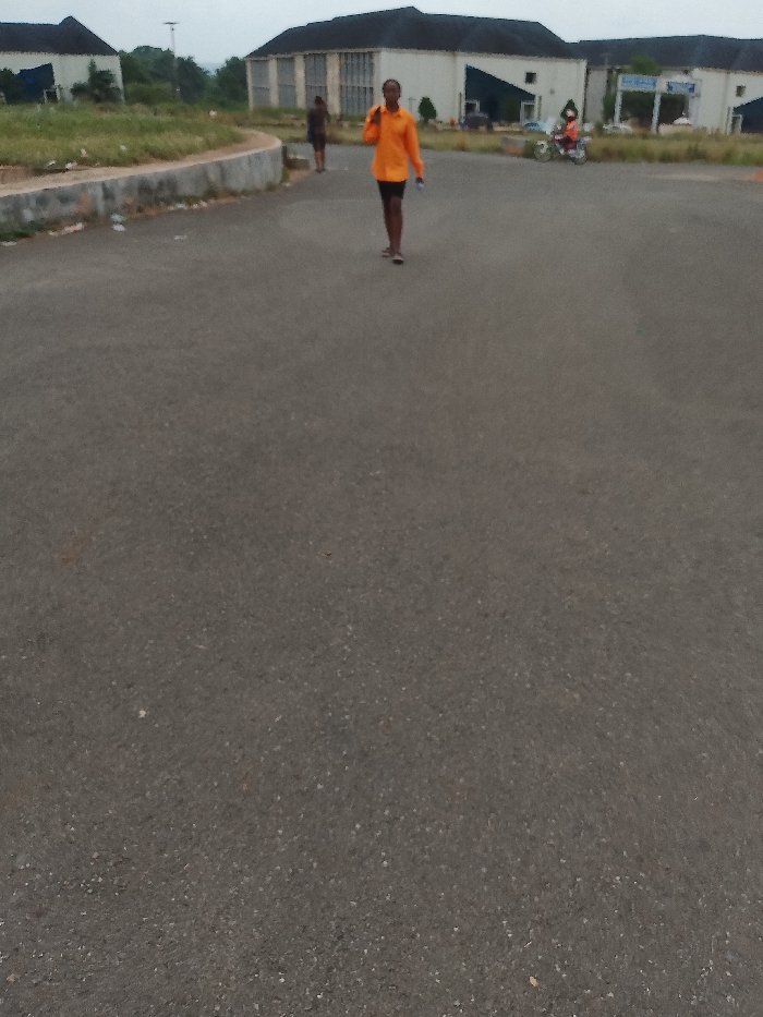 A paved road stretches toward a series of buildings in Ogboko, Nigeria. A person wearing an orange shirt and dark shorts is walking on the road, holding a phone to their ear. A second person is walking behind them to the left. A person on a motorcycle is visible in the background near one of the buildings. The buildings appear to be institutional or public structures. The sky is overcast. Debris and trash are scattered along the edge of the road.