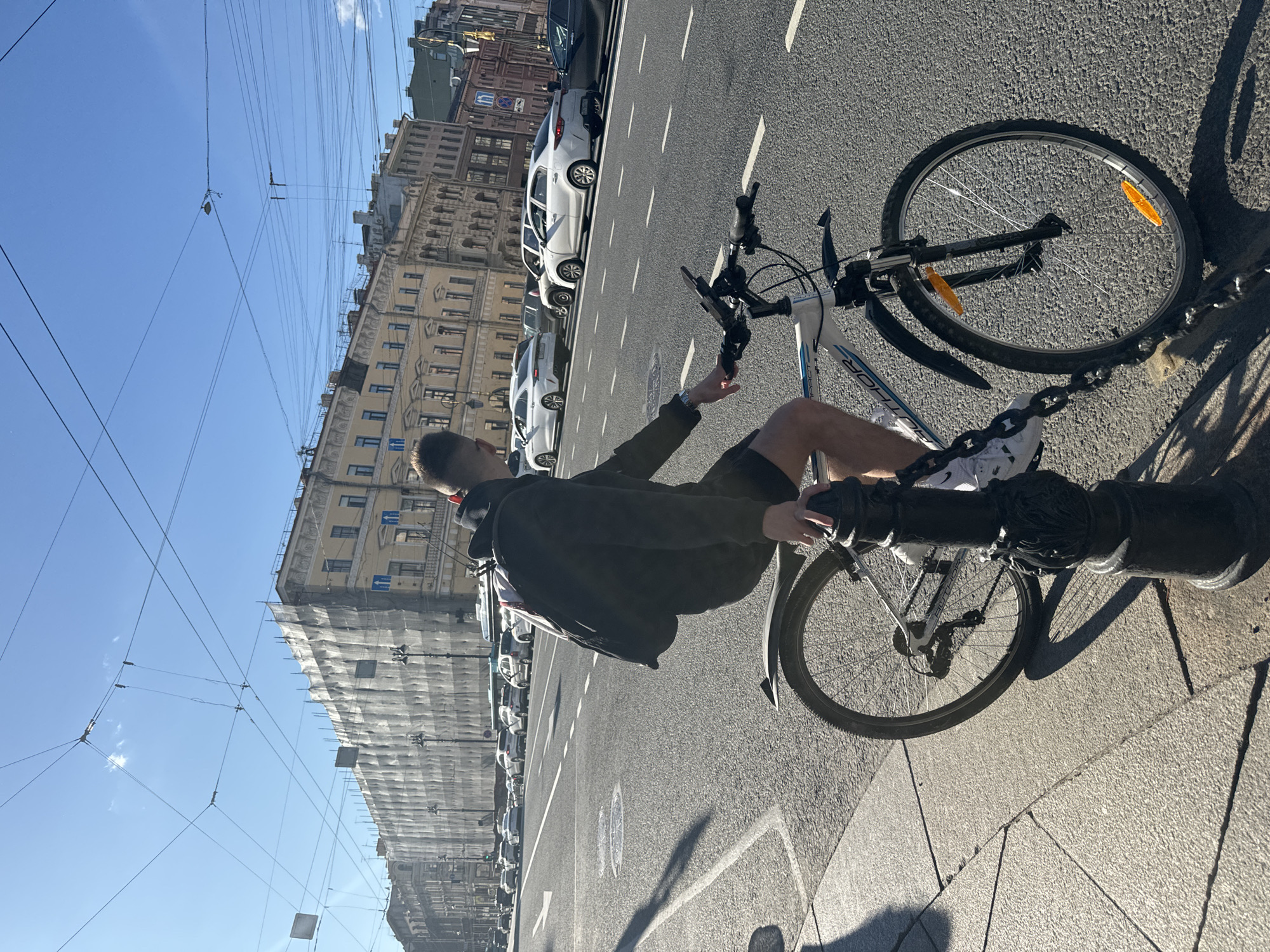 A young man with short hair, wearing a dark hooded jacket and black shorts, is seated on a bicycle in Saint Petersburg, Russia. He is positioned on the right side of the frame, facing away from the camera and towards the street. The bicycle is white and black with blue accents and is locked to a decorative black bollard with a chain. The man has a black backpack on his shoulders. The scene takes place on a sunny day with a clear blue sky and some scattered clouds. A wide street with multiple lanes of traffic is visible in the background, with several cars driving. On the left side of the street, an old European-style building is under renovation, covered in scaffolding and white sheeting. The architecture of the buildings suggests a historic urban environment, characteristic of Saint Petersburg. The pavement is made of stone slabs, and there are tram wires overhead, indicating this is likely a street with public transportation.