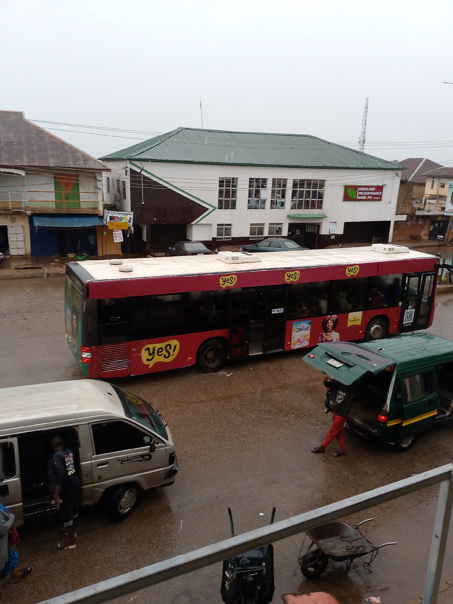 The image shows a street scene in Bukuru, Nigeria on a rainy day. A red and white "yes!"-branded bus is stopped on a wet road, with images of "Cornflakes" and "NASCO" along its side. A silver van is parked nearby with its door open, and a person is standing outside the van. A green van with an open trunk is also visible. A man in red pants is walking. A wheelbarrow and luggage are visible near the bottom of the frame. Buildings are visible in the background, including one with a sign for "HIGHLAND MICROFINANCE BANK Plc."