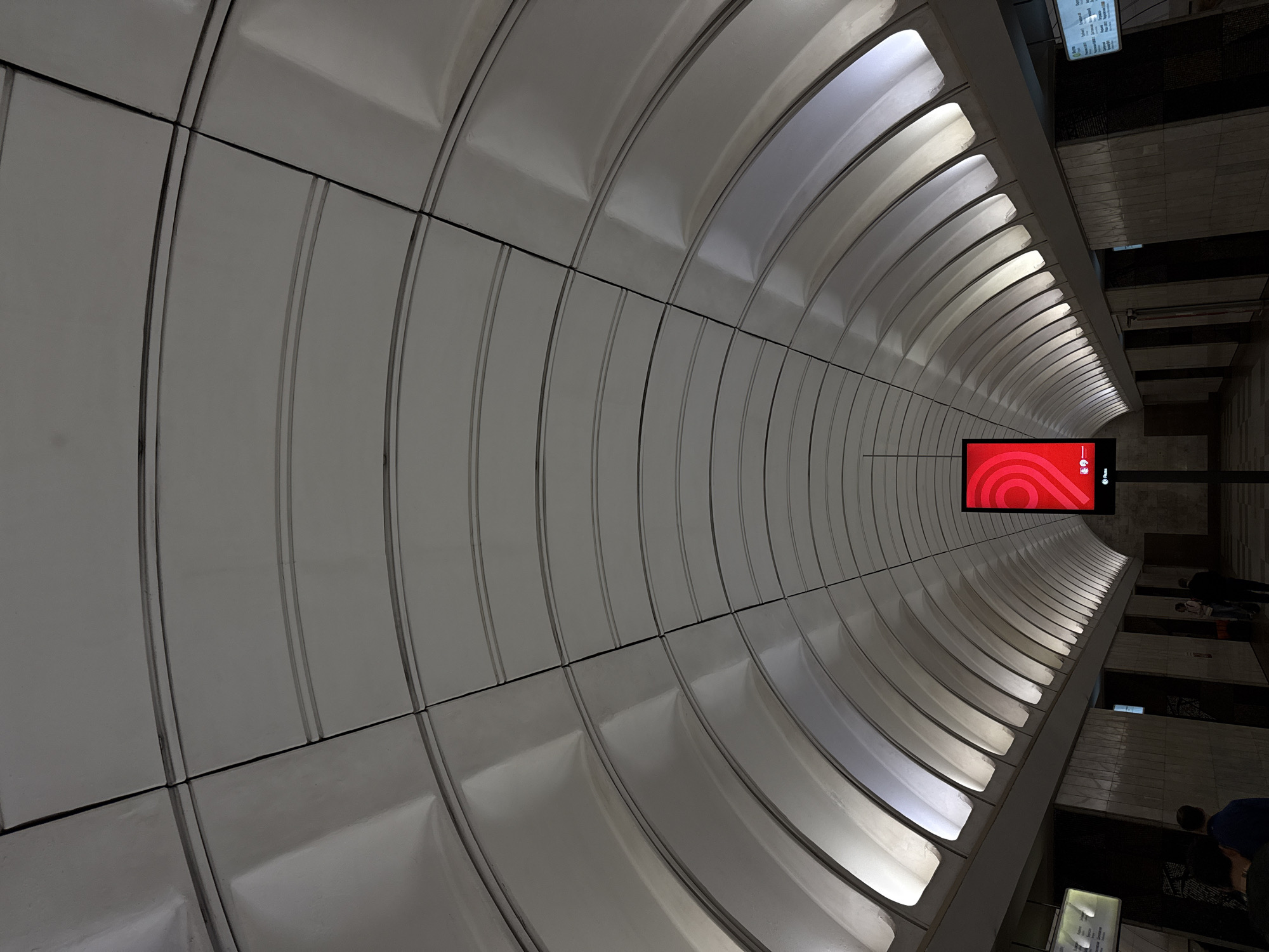 This is a wide-angle, low-angle shot of a Moscow Metro station ceiling. The ceiling is a ribbed, vaulted structure made of white panels, with recessed lighting along the edges of the ribs. A large, rectangular digital advertisement screen with a bright red background and a circular graphic is suspended from the ceiling in the middle distance. The station has tiled walls and a checkered floor. Several people are visible in the background, standing or walking. The image captures a sense of spaciousness and modern design typical of many Moscow Metro stations.
