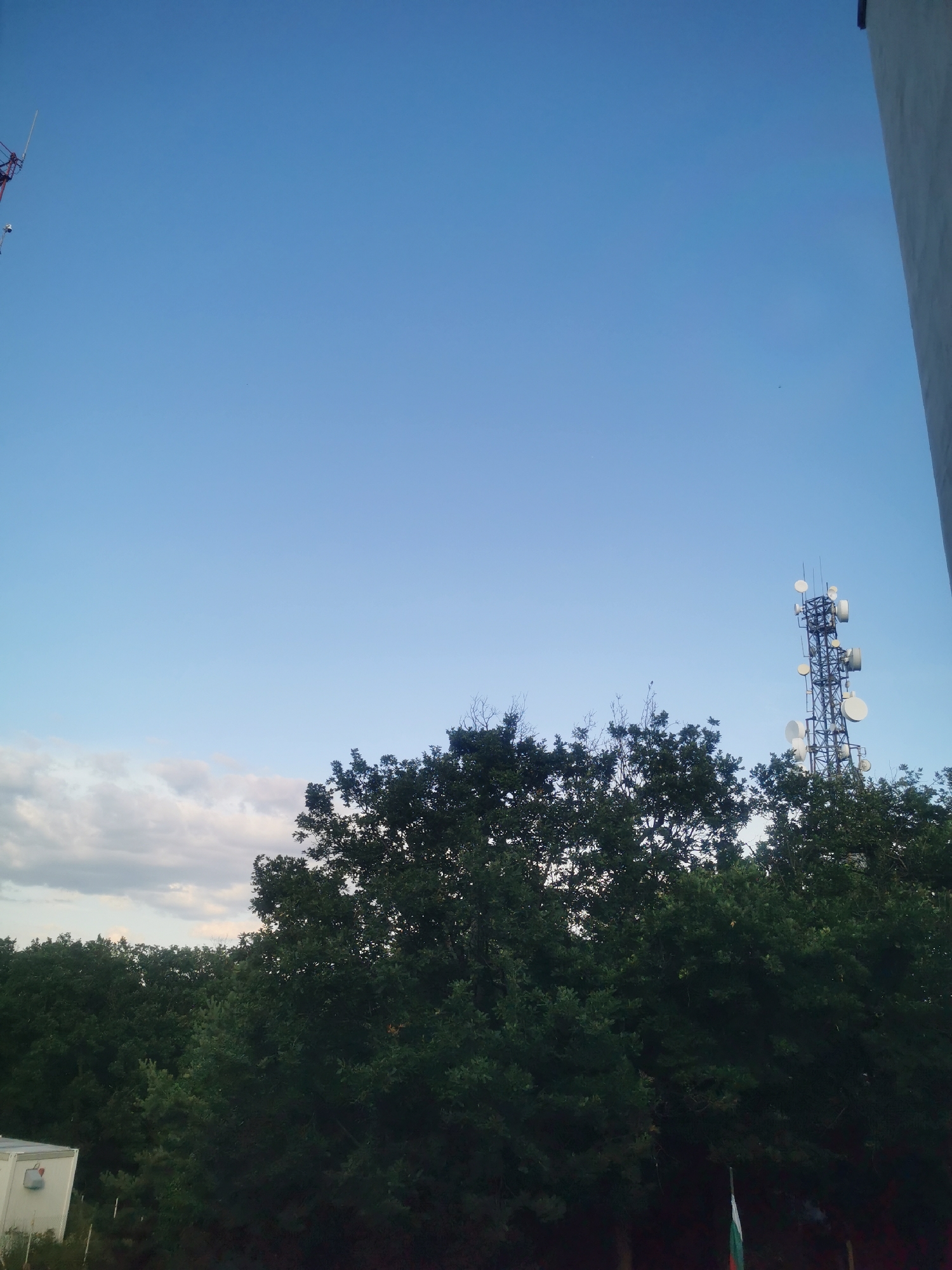 The image shows a clear blue sky above a dense canopy of green trees. In the upper left quadrant, the top of a red and white structure, possibly a crane or antenna, is visible. To the right, a tall, black communication tower with multiple antennas and dishes stands amidst the trees. On the far right edge, a sliver of a white building is present. In the lower left, a white prefabricated structure with a red electrical box is visible. In the lower right, a Bulgarian flag, partially obscured by trees, is flying. The scene appears to be an outdoor environment in Elhovo, Bulgaria, with a mix of natural and man-made structures.