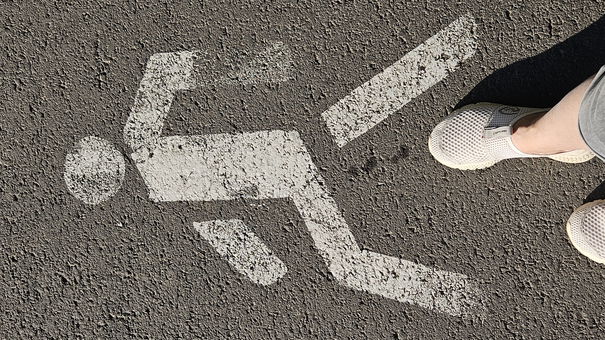 An overhead, high-angle shot captures a white pedestrian symbol painted on a grey asphalt surface. The symbol depicts a simplified human figure in motion, with a circular head and a body composed of rectangular and linear shapes suggesting walking. The asphalt is textured and shows signs of wear. In the foreground, two feet wearing light-colored mesh slip-on shoes are visible, casting a shadow to the right. The context suggests this is a pedestrian crossing or pathway, likely in Almaty, Kazakhstan, given the implied location.