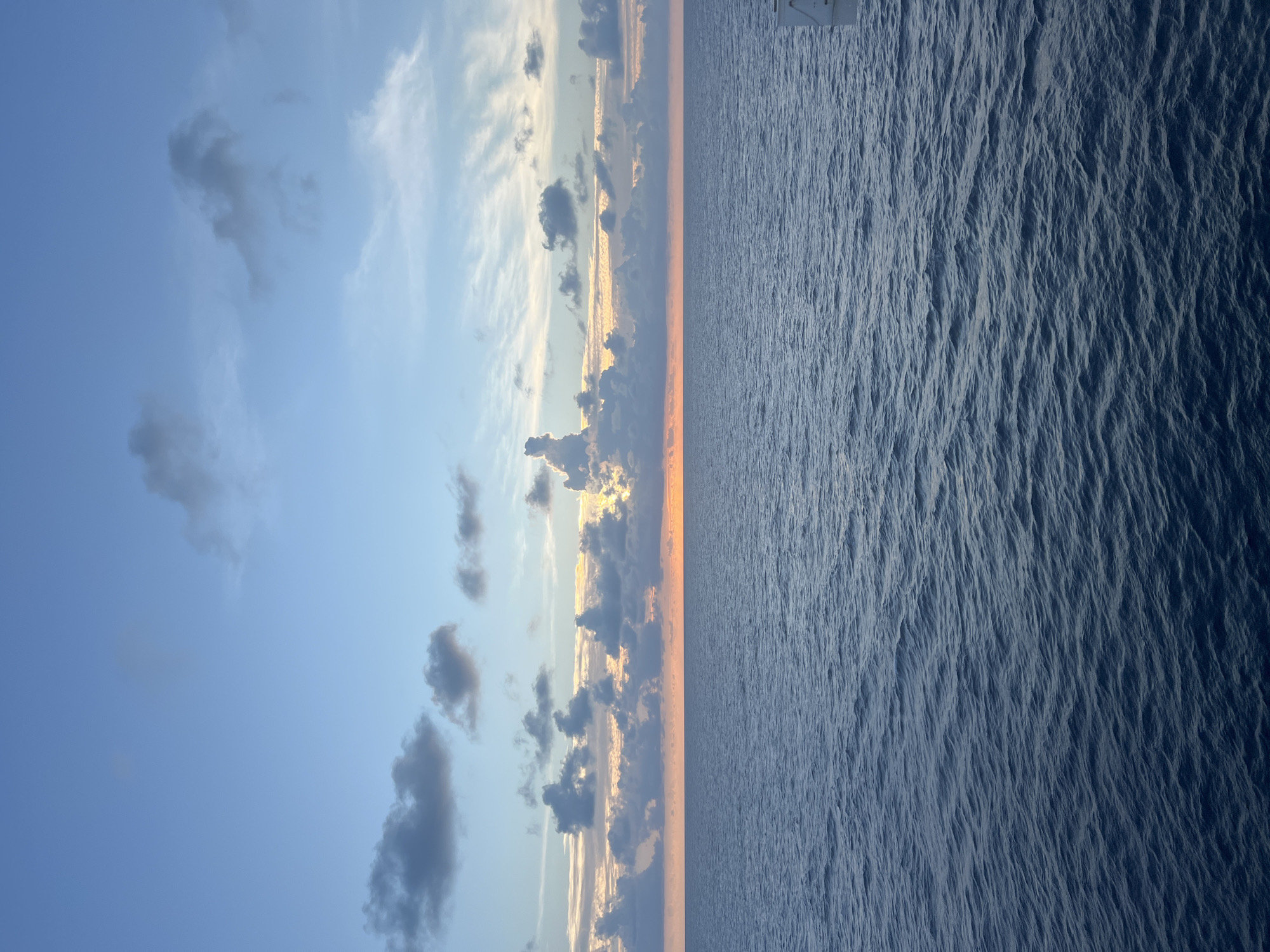 The image depicts a serene ocean sunset. The sky is a gradient of blue, transitioning to hues of orange and yellow near the horizon where the sun is setting. Scattered clouds, some dark and others wispy, dot the sky. The ocean below is calm, with gentle ripples reflecting the colors of the sky. The horizon line is sharp and clear. The lower right corner of the image shows a portion of a white structure, possibly part of a ship or a pier, extending into the frame. The scene is devoid of any people or explicit city elements, suggesting an open-water or coastal environment.