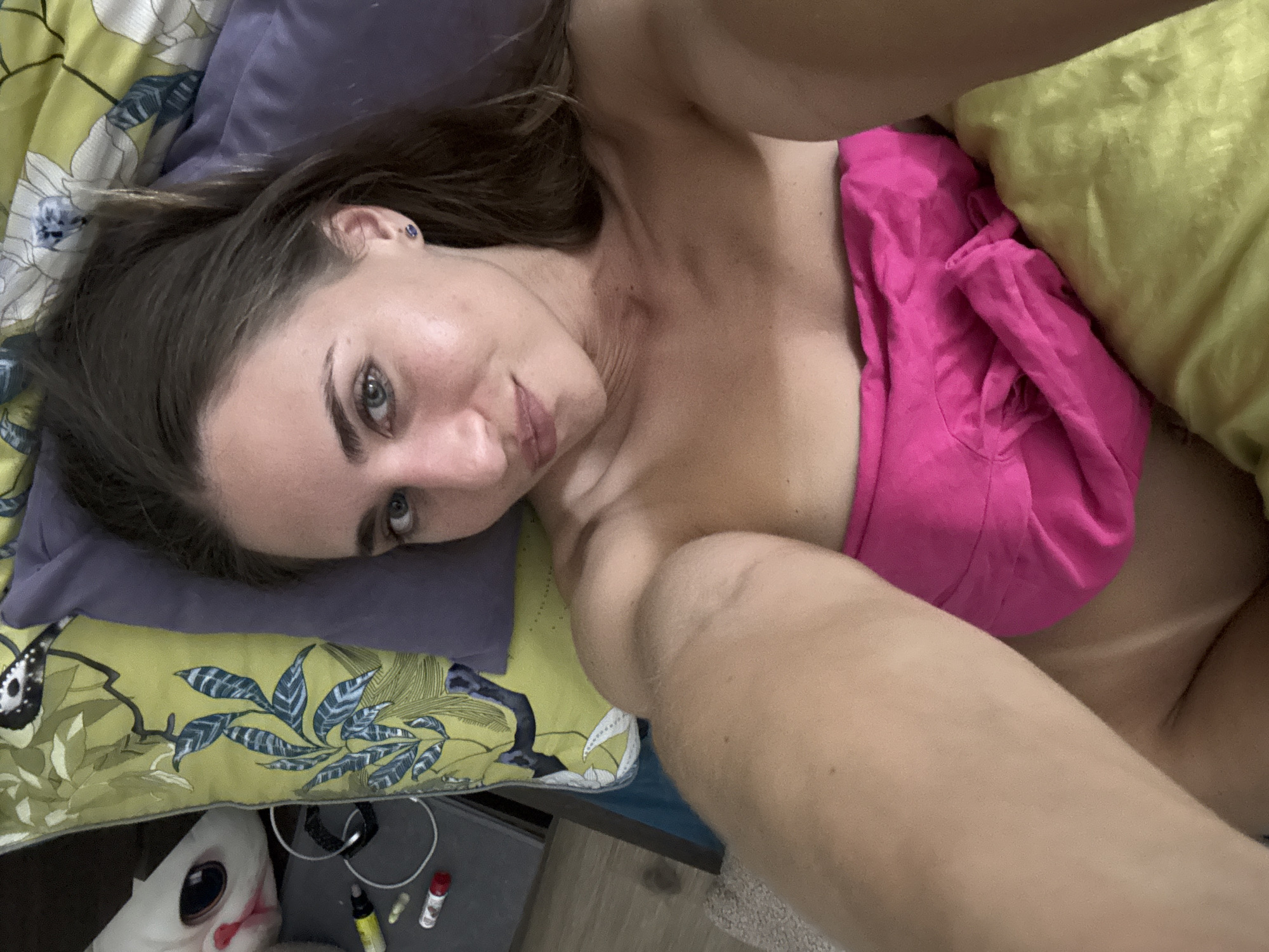 A woman is lying in bed, facing the camera, taking a selfie. She is covered from the chest down with a pink cloth. She has brown hair and blue eyes. She is wearing a small blue earring in her right ear. A stuffed animal and some pens are on the bed next to her. The room is decorated with a floral pattern on the pillow. The background appears to be a bed with yellow and purple pillows and a patterned comforter. The lighting is soft and natural.