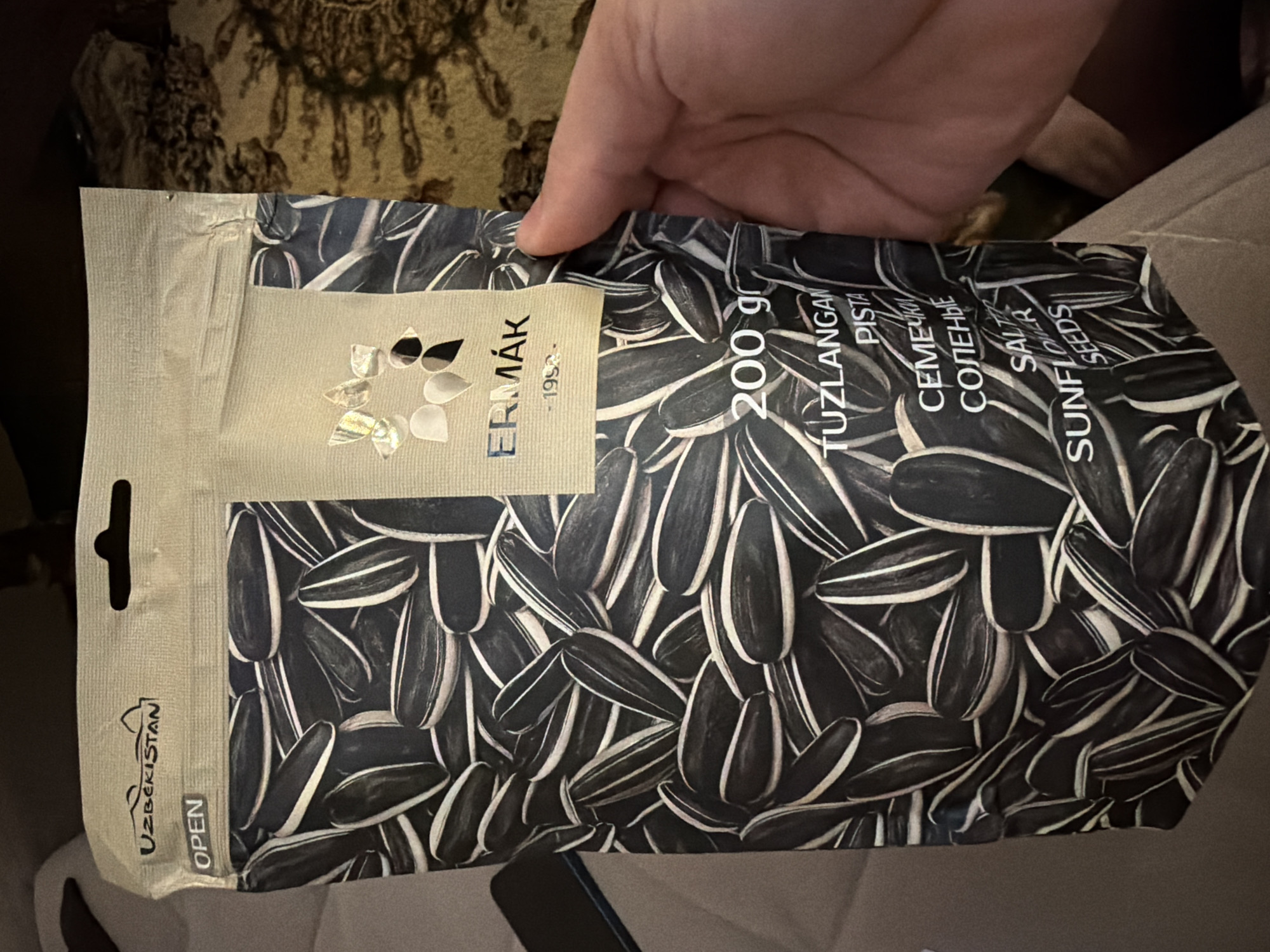 A person's hand is holding a bag of sunflower seeds. The bag is predominantly black and white, featuring a pattern of sunflower seeds. The bag has text and graphics printed on it. The brand name "ERMAK" is visible in silver lettering, along with "1952" underneath. "UZBEKISTAN" is also printed at the top of the bag. The weight "200 gr" is clearly displayed. Below "200 gr", text in a foreign language is visible, including "TUZLANGAN PISTA", "SEMECHKI", "SOLENYIE", "SALTED", and "SUNFLOWER SEEDS". The background of the image is blurred and appears to be an indoor setting with patterned fabric. There is no identifiable city of Koshkurgon, Uzbekistan in this image.