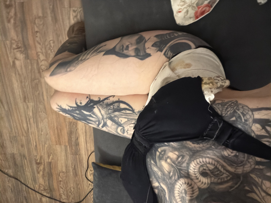 On a couch with pillows is a tattooed person wearing a black bra. The visible body parts include arms and the upper part of the torso, covered with intricate black and white tattoos of faces and serpentine figures. The person is lying down on the dark gray couch. One of the patterned pillows is white with floral accents. Part of a leg is visible at the top left. The background includes a wooden floor and a black cable.
