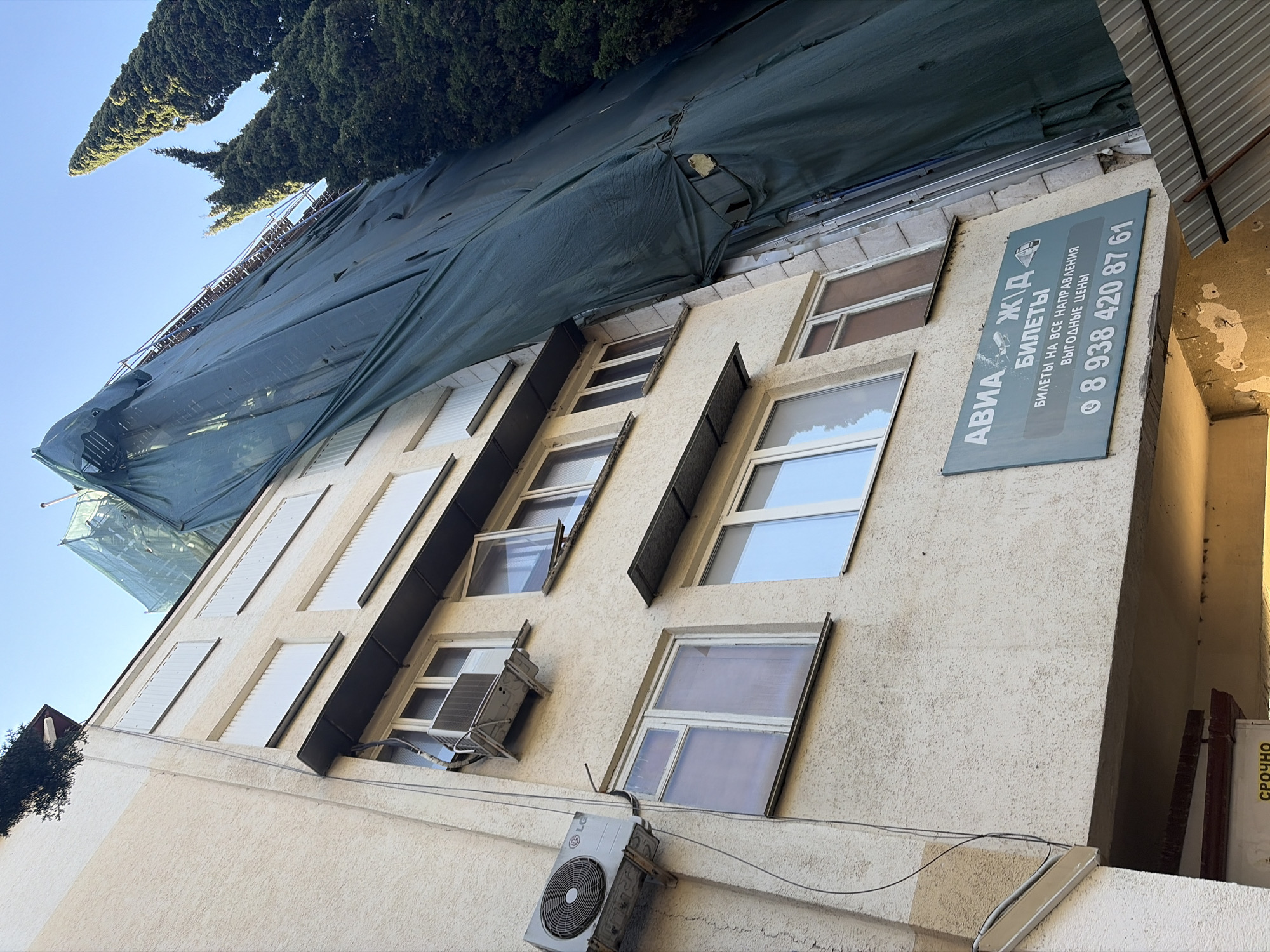 This is an upward-angled, outdoor photograph of a building facade in Sochi, Russia, captured under a clear blue sky. The upper section of the building is draped in a dark green tarpaulin, likely indicating construction, renovation, or scaffolding, with some structural elements visible beneath. Above the building, tall, dense evergreen trees (resembling cypress) are prominently featured against the sky.
The building's walls are a textured, light cream or beige color, punctuated by multiple windows. Some windows are equipped with white external blinds, while others appear to be partly open. Several air conditioning units are attached to the facade; a larger LG branded unit is mounted on the wall, and a smaller unit is installed within a window opening. Electrical wires run along the building's exterior.
A rectangular sign on the building displays travel-related information in Russian. The prominent text reads "АВИА Ж.Д. БИЛЕТЫ" (AVIA RAILWAY TICKETS), followed by "БИЛЕТЫ НА ВСЕ НАПРАВЛЕНИЯ" (TICKETS FOR ALL DIRECTIONS) and "ВЫГОДНЫЕ ЦЕНЫ" (FAVORABLE PRICES). A phone number, "8 938 420 87 61", is also clearly displayed. Additionally, partially visible text on a yellow background to the right of the building reads "СРОЧНО" (URGENTLY/EXPRESS).
The bright sunlight suggests it is daytime, either morning or afternoon, casting distinct shadows. No people are visible, and the scene depicts a static urban environment featuring architecture and natural elements.