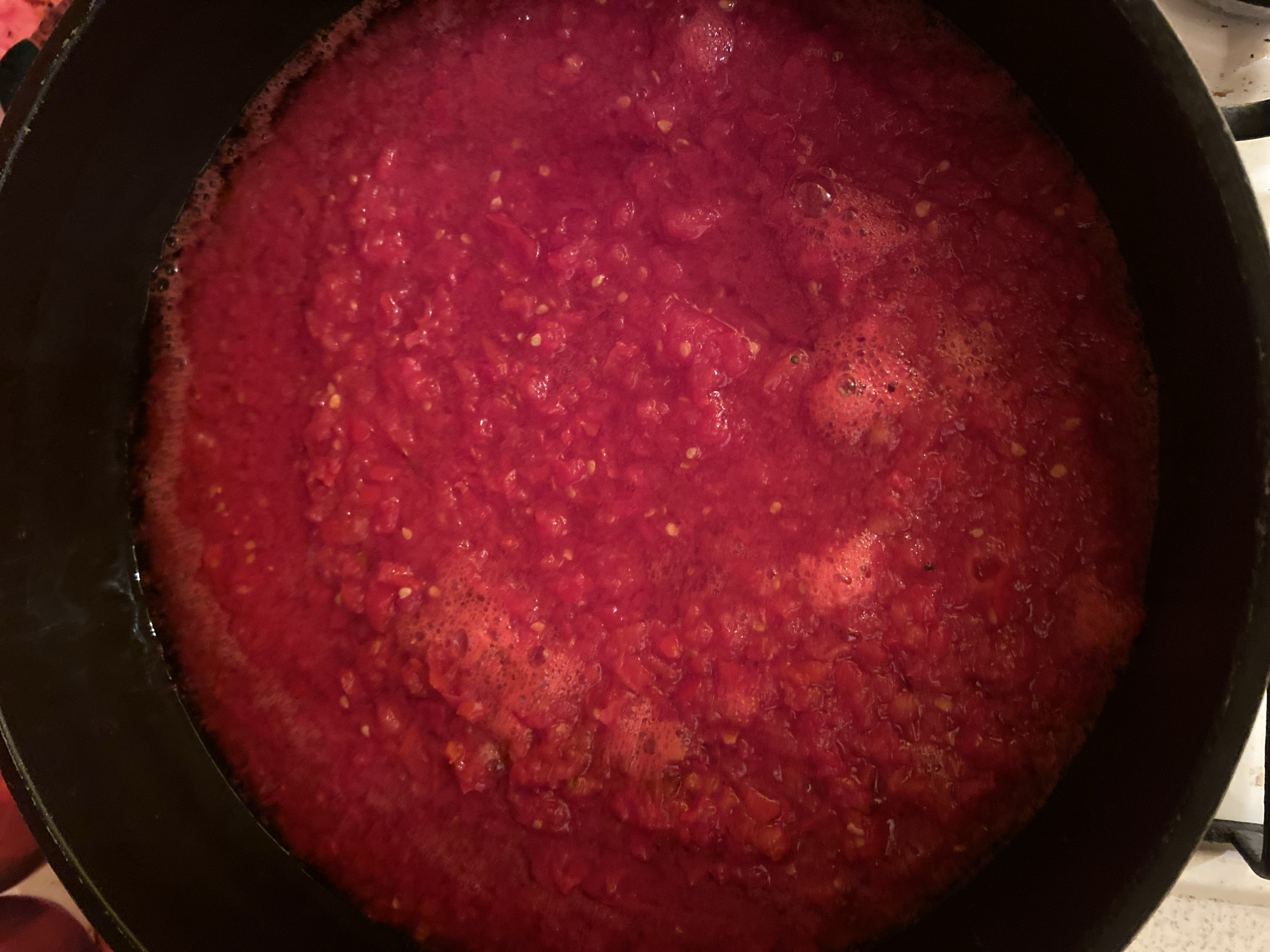 A close-up, top-down shot shows a large pot filled with a vibrant, red, chunky tomato sauce that is simmering. The sauce has a foamy texture on the surface and many small, visible seeds from the tomatoes. The dark rim of the pot is visible along the top and left edges of the frame, and a small section of a white stovetop is present at the bottom. The scene appears to be in a kitchen in Ivanteyevka, Russia.