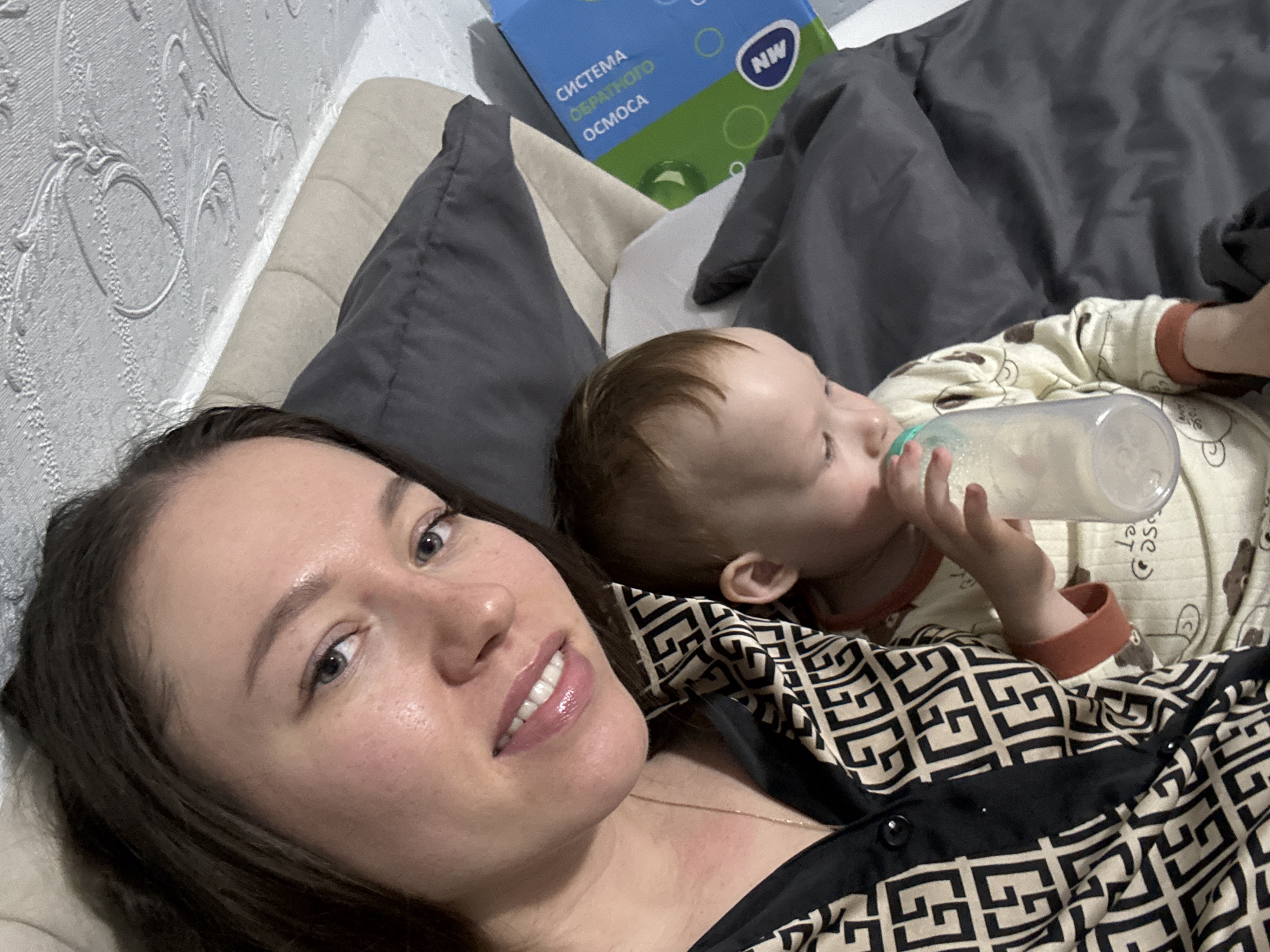 The image shows a woman lying on a bed next to a young child drinking from a baby bottle. The woman is in the foreground, facing the camera. She has dark hair and light skin. She is wearing a patterned black and beige shirt. The child is lying next to her, partially obscured by the woman's shoulder. The child is wearing a patterned beige onesie with brown cuffs. A baby bottle is visible in the child's hands and mouth. Behind the woman and child, there are pillows and a blue box with Russian text. The wallpaper is patterned. The location is Ферма, Russia.