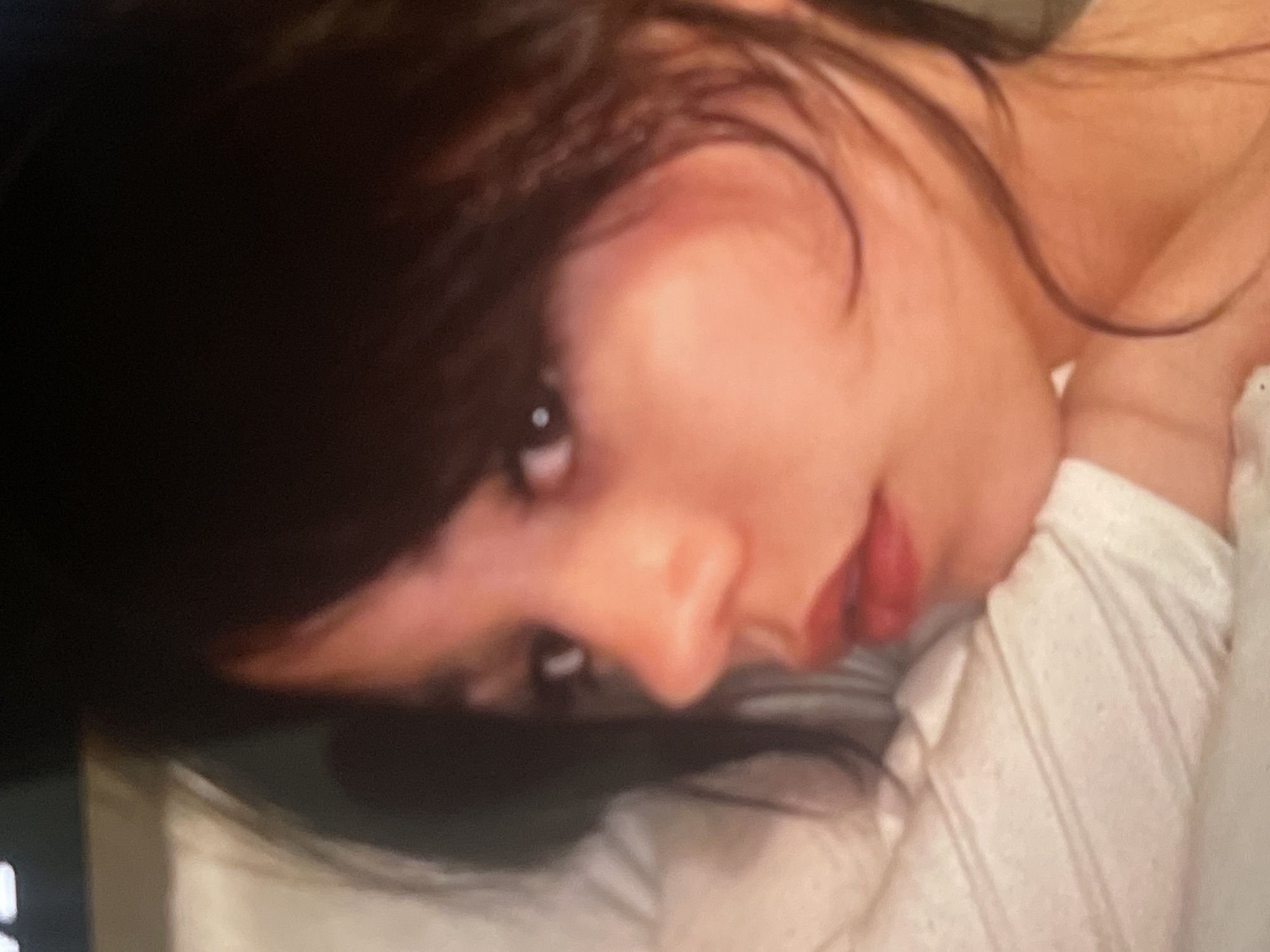 This close-up portrait features a young person, likely female, with dark hair, positioned in an indoor setting. The subject's head is tilted at an angle, and she looks directly towards the viewer with dark eyes. Her dark hair frames her face, with strands falling across her forehead and cheek. She has a defined nose and slightly parted lips, revealing a hint of natural color.
She is wearing a light-colored, possibly white, top with a discernible ribbed or textured pattern on the sleeve visible near her right shoulder. A faint, lighter strap or fabric edge is visible near her left shoulder/neck area. The background is dark and out of focus, suggesting an intimate, indoor environment with subdued or controlled lighting.
The person's expression appears calm and direct, conveying a sense of gentle engagement or introspection. There are no specific objects or visible text in the background. While the provided context indicates the location is Poltava, Ukraine, the image itself does not contain any specific visual cues to confirm this geographical location. The activity is clearly posing for a photograph.