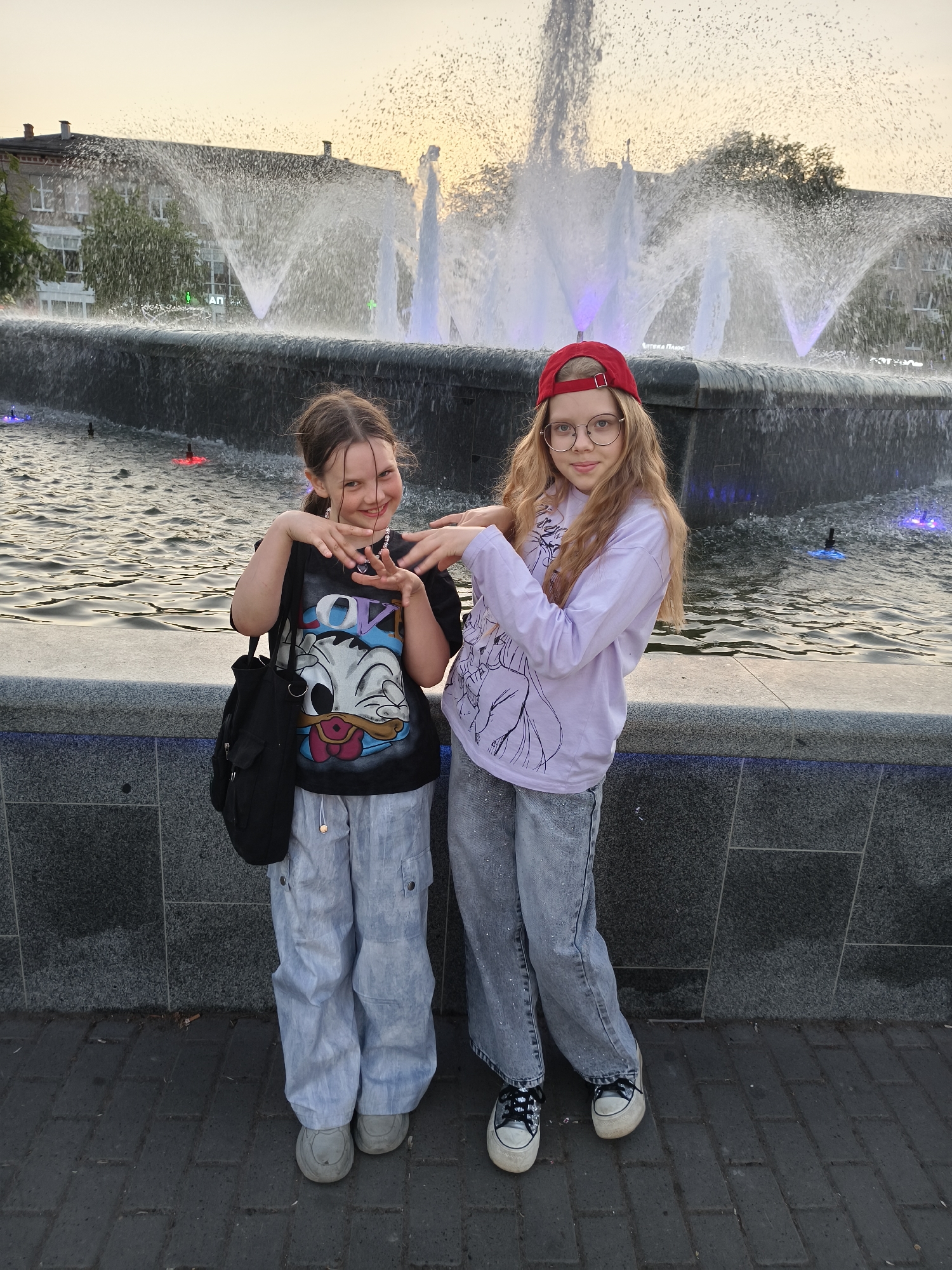 Two young girls stand in front of a water fountain in an urban environment. The girl on the left is wearing a black t-shirt with a graphic, baggy blue pants, and gray shoes. She carries a black tote bag on her left shoulder. The girl on the right wears a light purple long-sleeved shirt, blue jeans with sparkles, white sneakers, and a red cap, and she wears glasses. Both girls are posing with their hands in front of them. A large fountain with multiple streams of water stands behind them. The setting appears to be a public area in a city, with a building visible in the background. The ground is paved with gray bricks. The sky is light, indicating late afternoon or early evening.