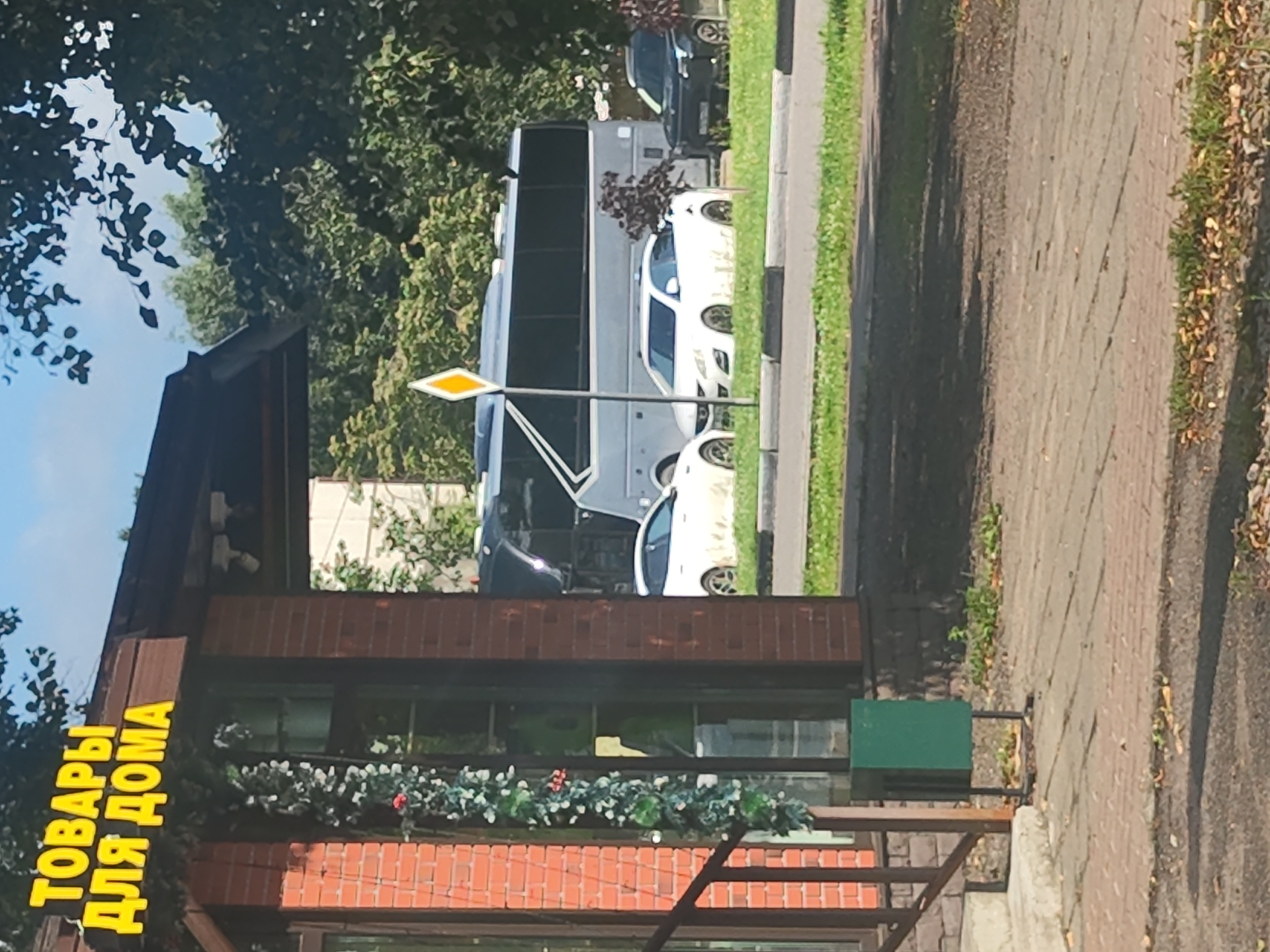 The image shows a street scene in Ramenskoye, Russia, on a sunny day with a clear sky. In the foreground, a brick building with a wooden overhang displays a bright yellow sign with the Russian text "ТОВАРЫ ДЛЯ ДОМА" (Goods for Home). To the left of the building, a metal railing leads up to a staircase. A green trash bin stands on the sidewalk.
Behind the building, a large gray bus is parked on the street. Several cars are visible in the background, including a white sedan parked on the grass near the bus, and a dark-colored SUV further to the right. A yellow diamond-shaped traffic sign is positioned on a pole between the bus and the white car. Trees and foliage are present in the background, suggesting a suburban or urban area with greenery. The ground is paved with brickwork and asphalt, with patches of grass and fallen leaves.