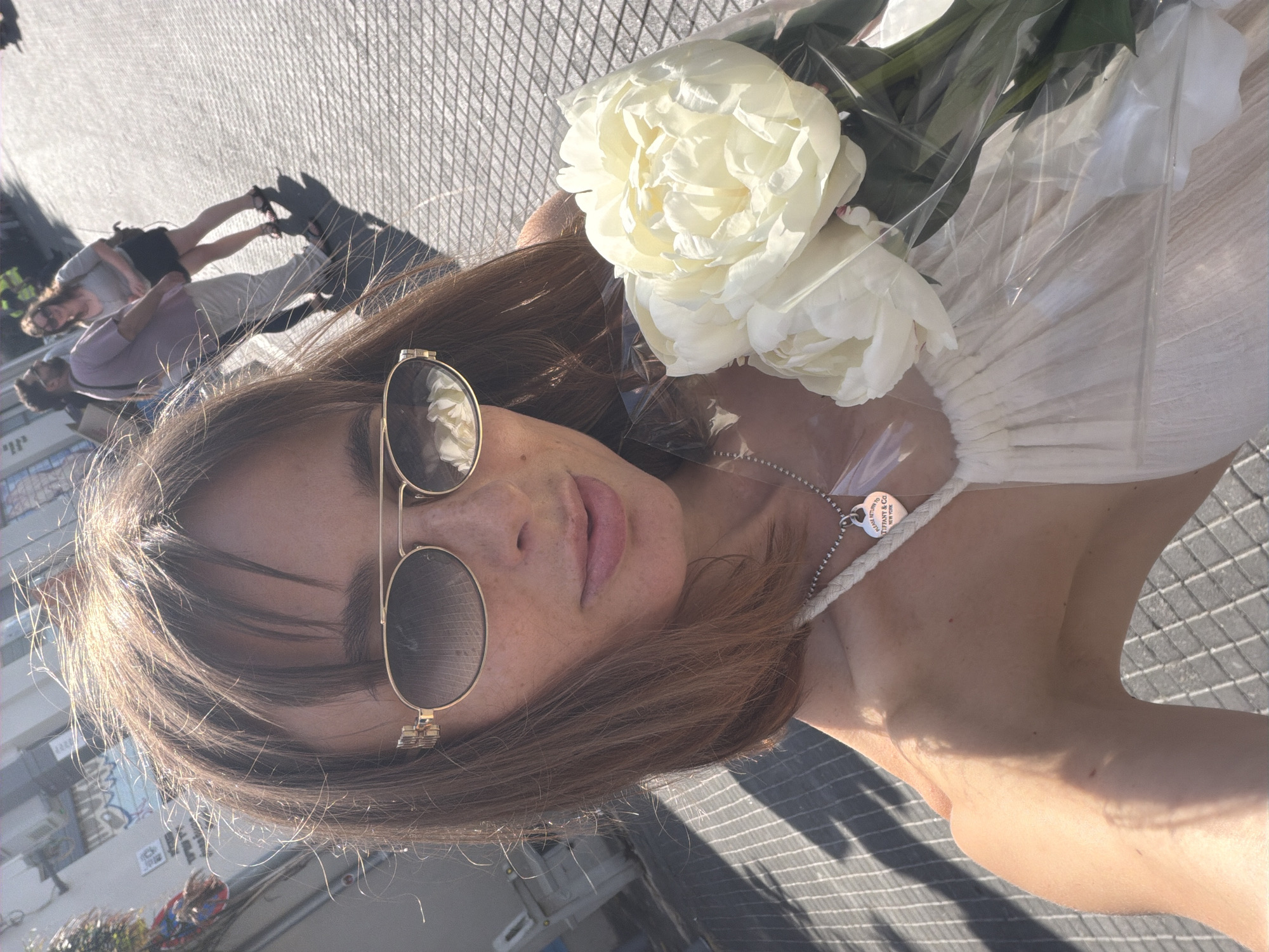 The original image is a selfie taken by a woman in Tel Aviv, Israel. The woman is wearing a white dress and sunglasses, and she is holding a bouquet of white flowers. In the background, there are other people walking on the street, and there are buildings and graffiti on the wall. The sun is shining, and there are shadows on the ground. The overall impression is a sunny day in a city.
The photograph shows a young woman with light brown hair, parted in the middle, and bangs. She is wearing oversized, oval-shaped sunglasses with thin gold frames. A small silver necklace with a heart-shaped pendant is visible around her neck. She is holding a bouquet of large, pale yellow peonies wrapped in clear plastic. The woman is wearing a light-colored, possibly white, dress with thin straps. Her skin is fair with freckles.
The background suggests an urban environment. The ground is paved with a textured pattern, possibly cobblestones or interlocking pavers, with distinct linear shadows indicating sunlight. Behind the woman, a blurry background shows other individuals walking, one in a light purple top and beige pants, and another woman in a grey top and dark skirt. There is also graffiti visible on a wall on the left side of the image. The presence of these elements, combined with the distinctive paving, points towards a location in Tel Aviv, Israel. The general lighting and attire suggest a warm, daytime setting.