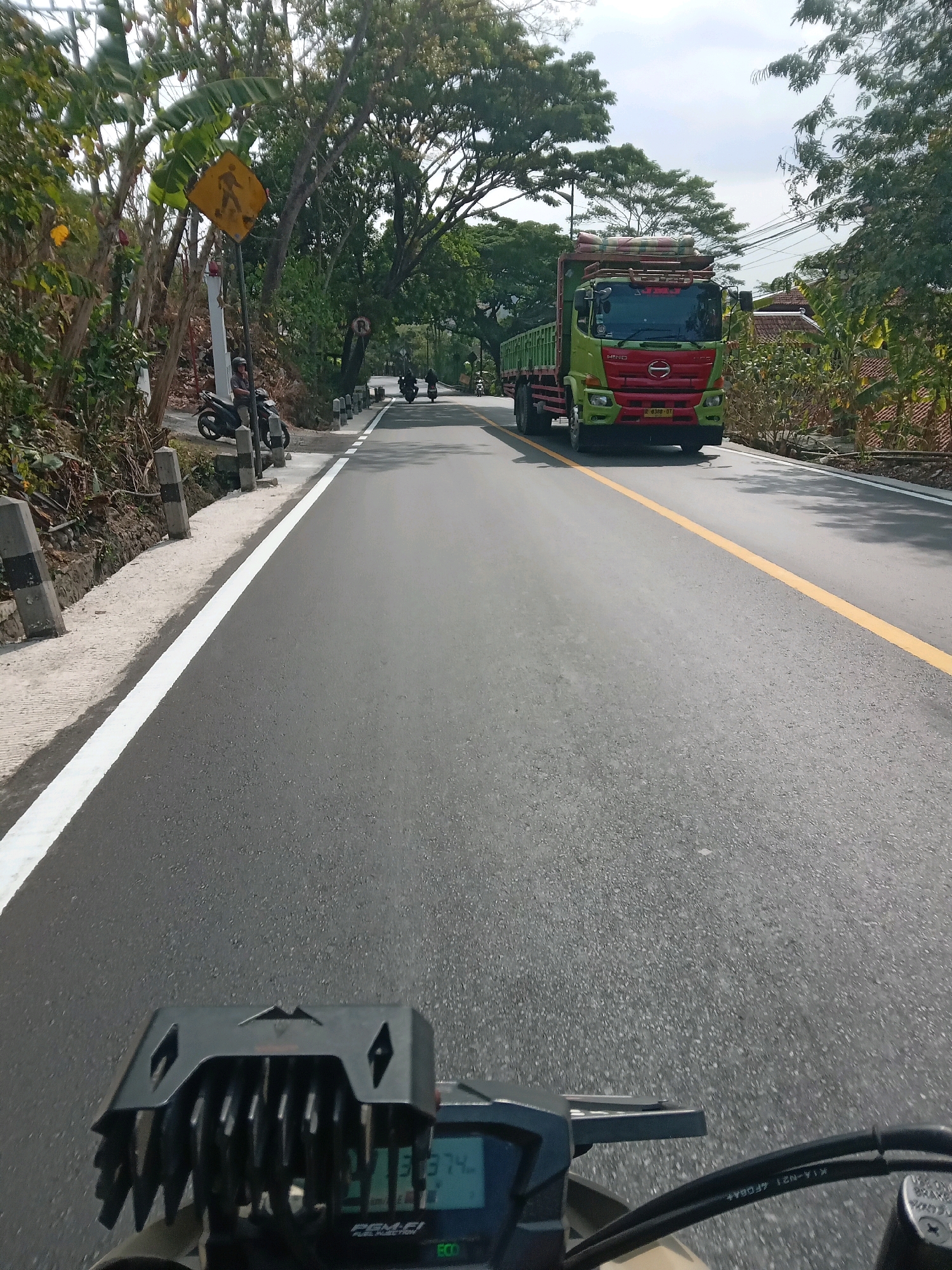 The image captures a view from a motorcycle on a paved road in Srimartani, Indonesia. The road has a yellow center line and a white line along the edge of the asphalt. To the left of the road, there is a sidewalk with concrete posts and a yellow pedestrian crossing sign. Trees line the left side of the road, and a single motorcycle is parked on the side. In the distance, a red and green truck is driving on the road, and several other motorcycles are also visible on the road ahead. The background consists of lush green trees and some houses. The sky is overcast. The motorcycle in the foreground has a digital display showing speed and other indicators.