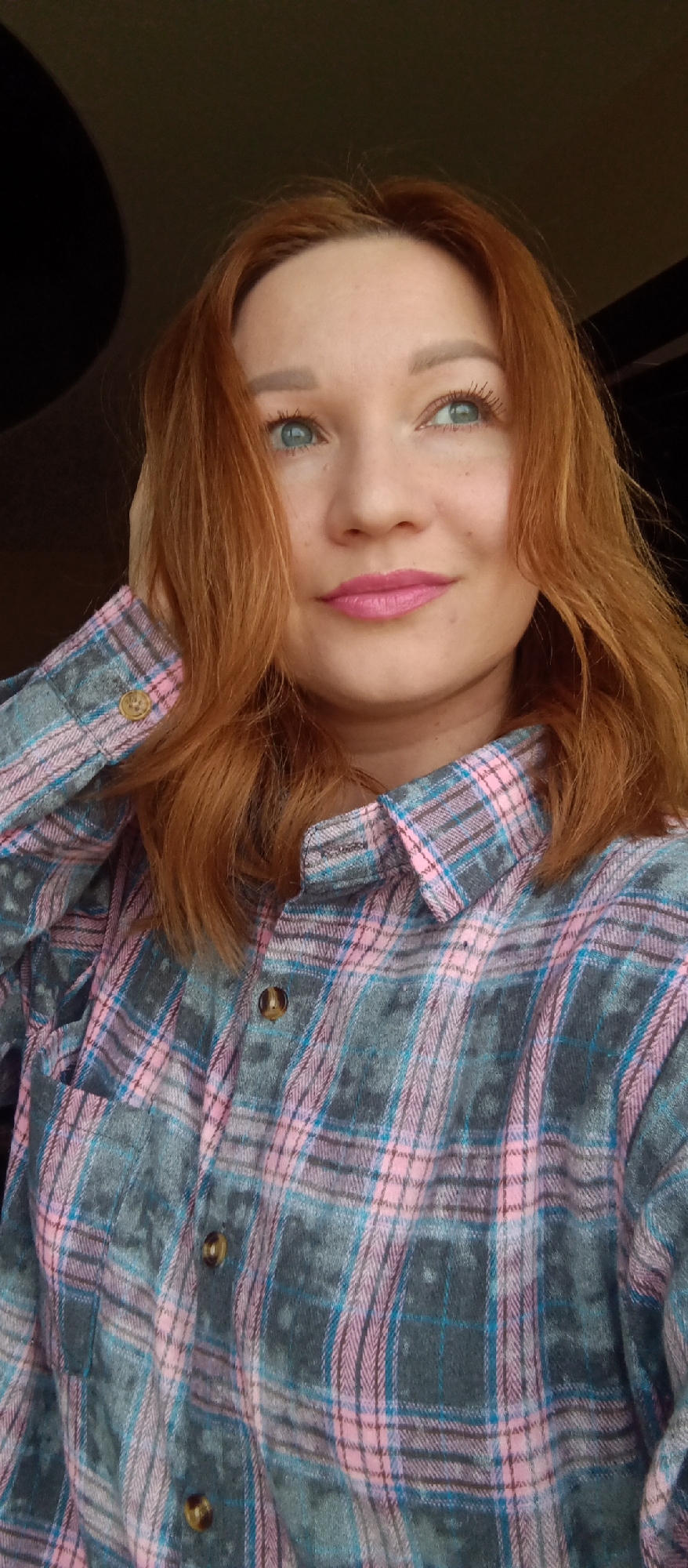 A close-up, chest-up shot features a woman with reddish-brown, wavy hair and striking blue-green eyes. She is looking upwards and slightly to the right, with a soft, subtle smile on her lips, which are colored with bright pink lipstick. Her right hand is gently raised, with her fingers touching her hair near her temple.
She is wearing a collared, long-sleeved plaid shirt with a pattern of grey, pink, and light blue squares. The shirt appears to be made of a soft, possibly flannel, fabric, and has dark brown buttons on the placket and sleeve cuff. Her eyebrows are neatly shaped, and she is wearing mascara. The background is dark and indistinct, offering no specific location cues. The lighting suggests an indoor setting or a shaded outdoor area. No visible text is present in the image.