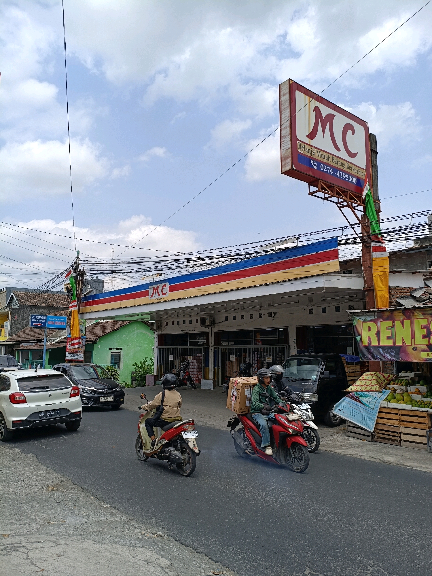 The image depicts a street scene in Maguwoharjo, Indonesia, dominated by a convenience store, branded "MC," with a prominent red, yellow, and blue striped awning. A large red and white sign for "MC" is mounted on a pole above the store, featuring text that reads "Belanja Murah Barang Berkualitas" and a phone number "0274-4395300."
In the foreground, two people are riding motorcycles on a paved road. The person in front is operating a red motorcycle and is wearing a green jacket and blue jeans. A large cardboard box is secured to the back of their motorcycle. The rider behind them is on a lighter-colored motorcycle and is wearing a tan jacket. Both riders are wearing helmets.
To the left of the store, a white car with license plate "AB 1495 NP" is parked or driving on the road. To the right of the store, a fruit stand is visible, displaying various fruits in wooden crates. The environment appears to be an urban or semi-urban setting with a clear sky and scattered clouds. Overhead, numerous electrical wires are strung across the scene. A green building is situated to the left of the "MC" store. A sign for "Dr. Wahyudi" and "PRAKTEK DOKTER" is also visible on the left side of the image.