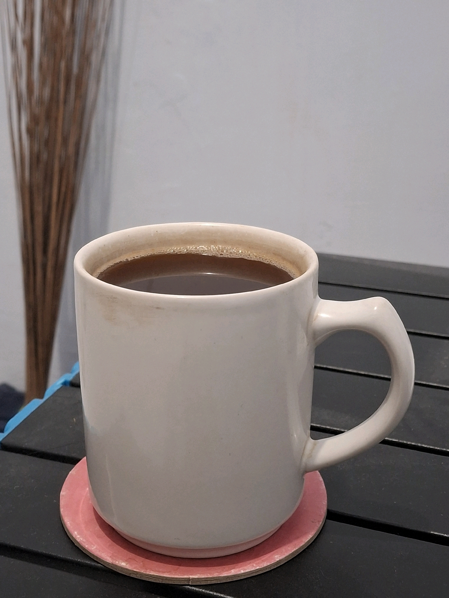 A close-up, eye-level shot shows a white ceramic mug filled with dark coffee, sitting on a pink coaster. The mug is placed on a black, slatted surface, likely a table. The coffee has a slight froth on top. To the left of the mug, a blurred object resembling a bundle of dried reeds or branches is visible against a light gray background. The scene appears to be indoors, possibly in Lumajang, Indonesia, given the context of the query, but there are no specific visual cues within the image to confirm this location. The lighting is soft and diffused.